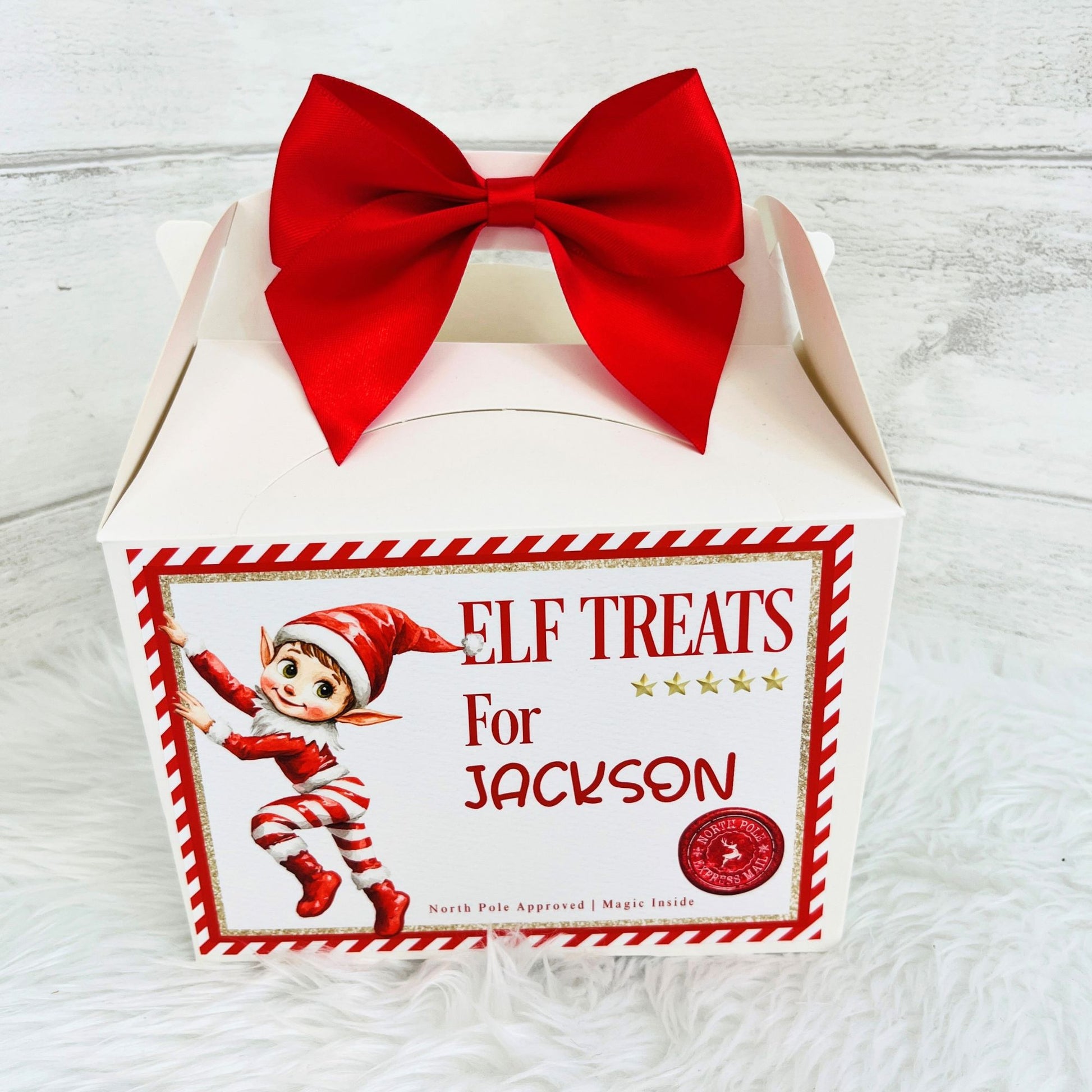 A white gift box with a large red bow on top, featuring a front label that reads 'ELF TREATS For Jackson' with an elf illustration and a red circular seal. The box is photographed on a white faux-fur surface against a light wood background.