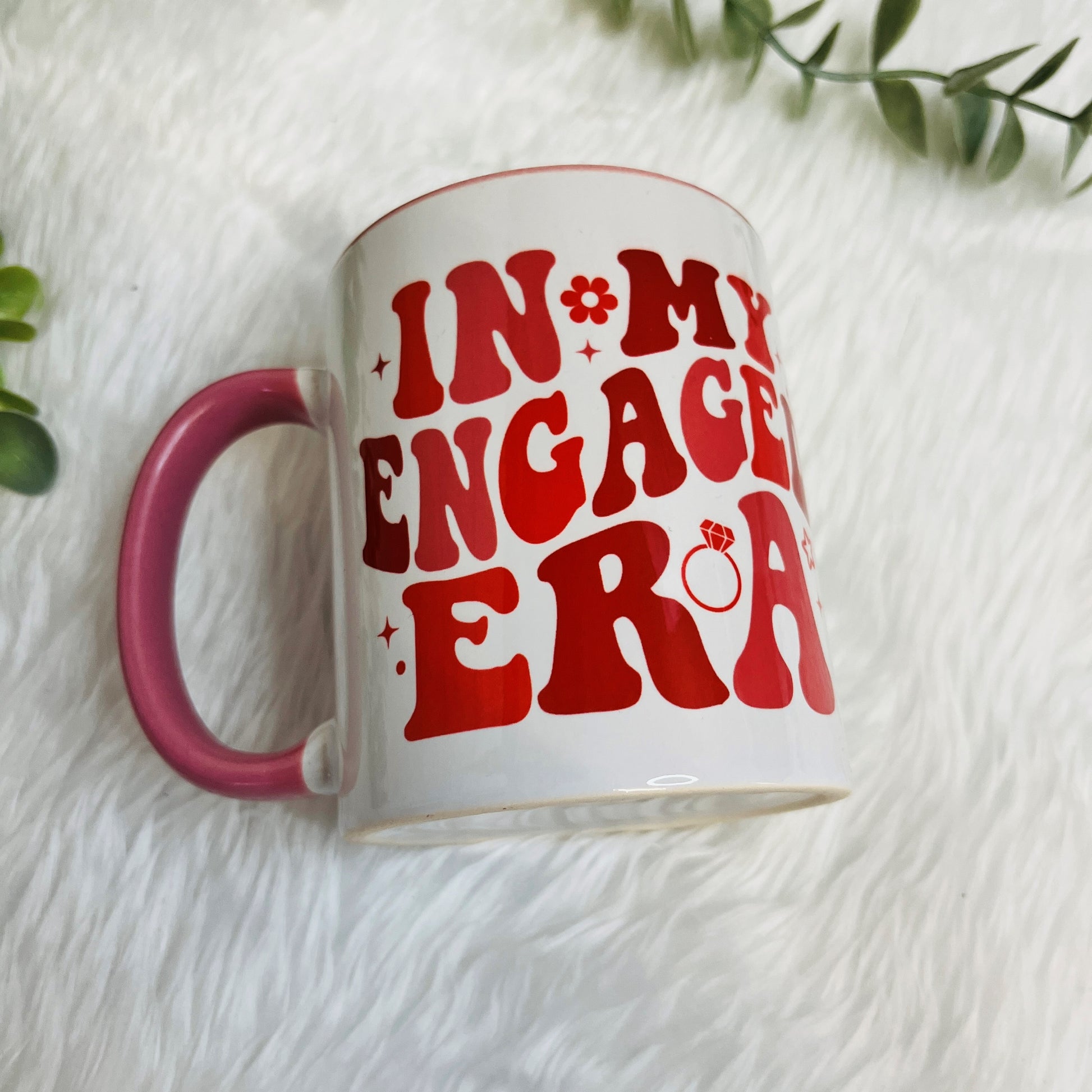 White mug with red text 'IN MY ENGAGED ERA' on a white surface with green leaves.