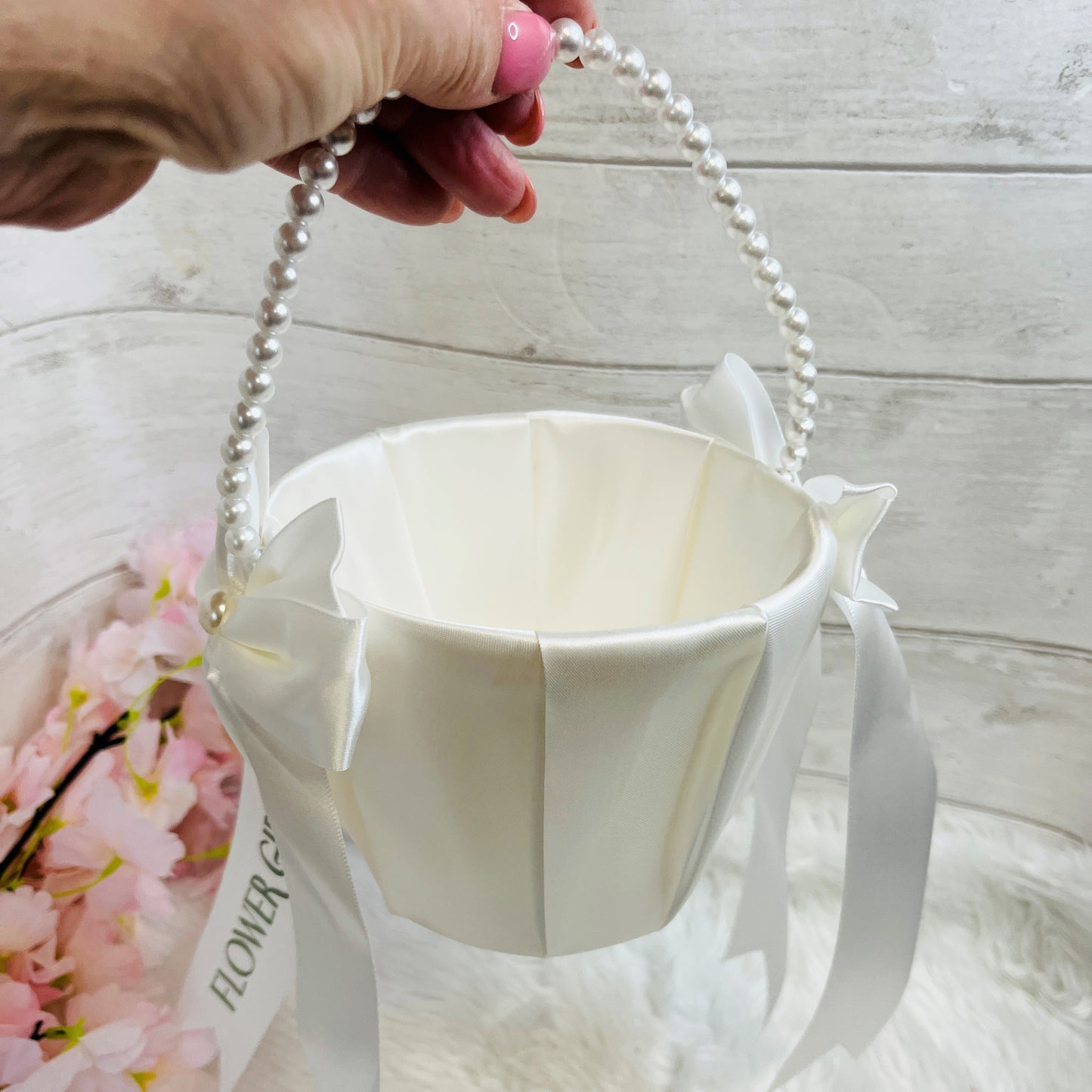 White flower girl basket with pearl handle held by a hand on a light wooden surface.