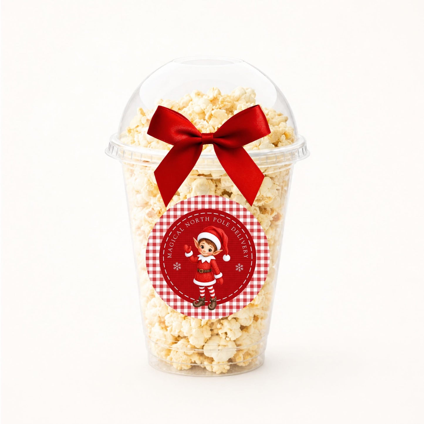A clear plastic popcorn cup filled with popcorn and topped with a large red ribbon bow, featuring a round red-and-white christmas elf sticker on the front.