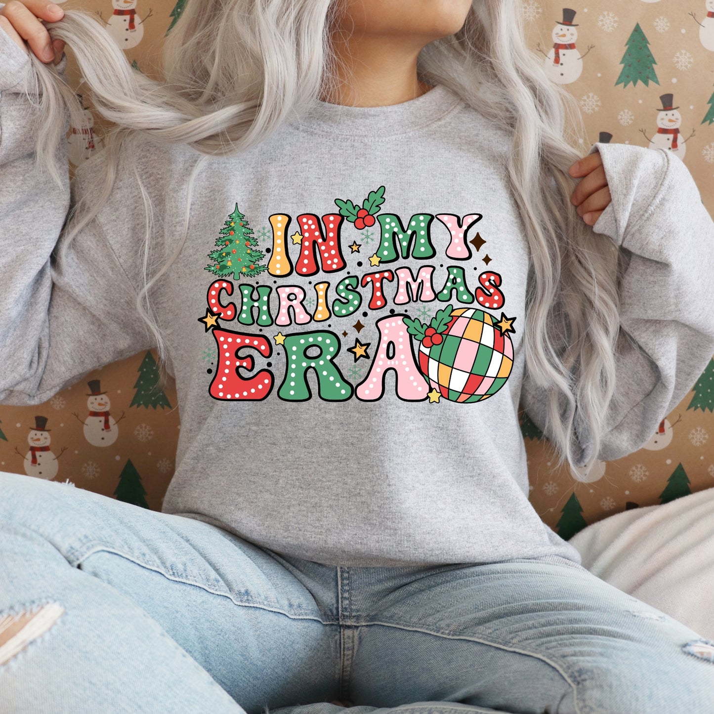 A model sitting with a grey jumper with 'In my Christmas Era' bright colourful design on the front