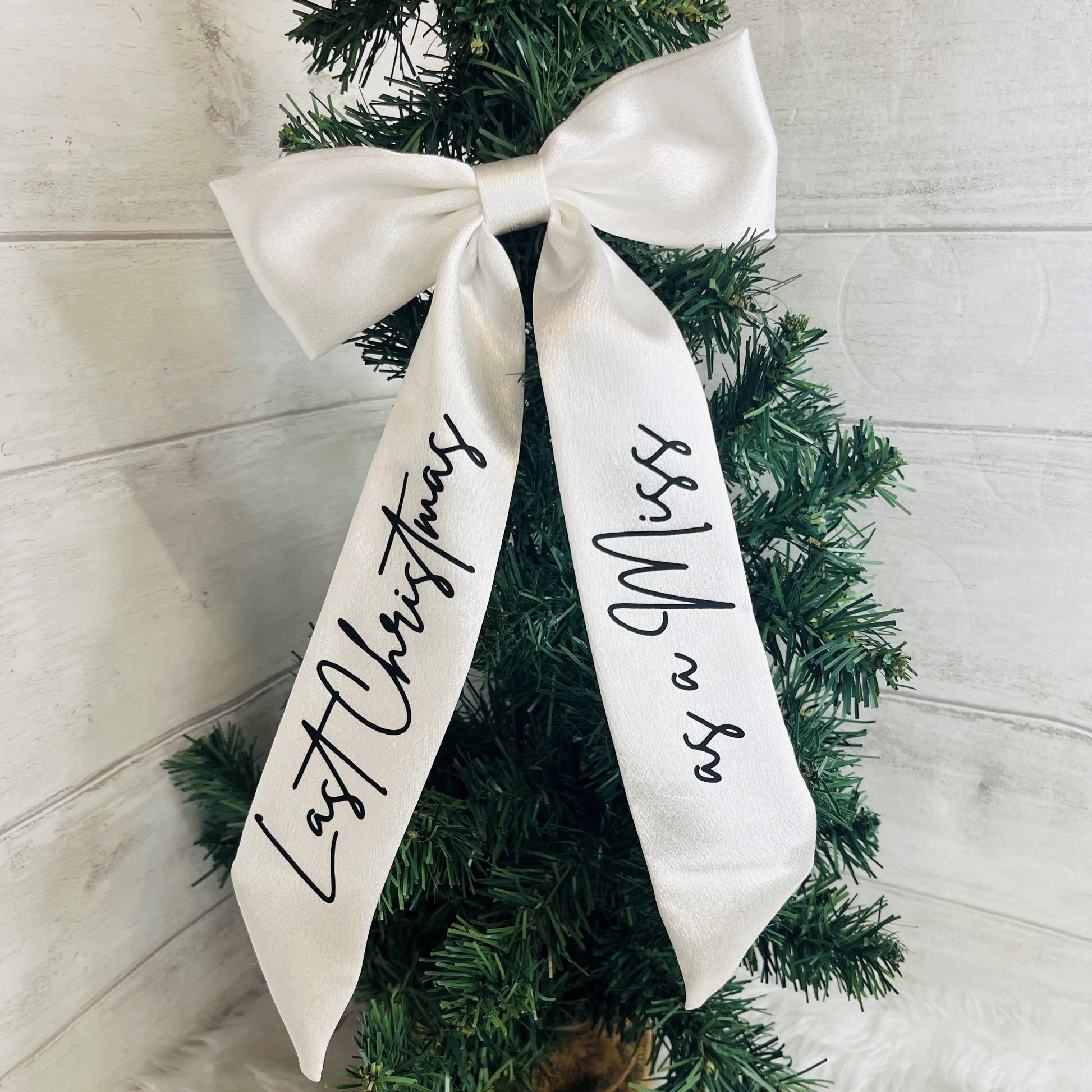 A white satin bow with black script text reading 'Last Christmas' on one ribbon end and 'as a Miss' on the other, tied to green artificial tree branches against a white wood plank background.