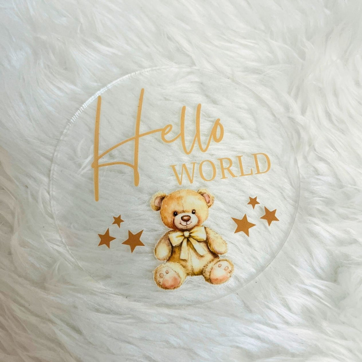 A round clear glass or acrylic coaster featuring a printed design of a cute teddy bear, gold stars, and the words 'Hello WORLD' in gold lettering, placed on a white faux fur surface.