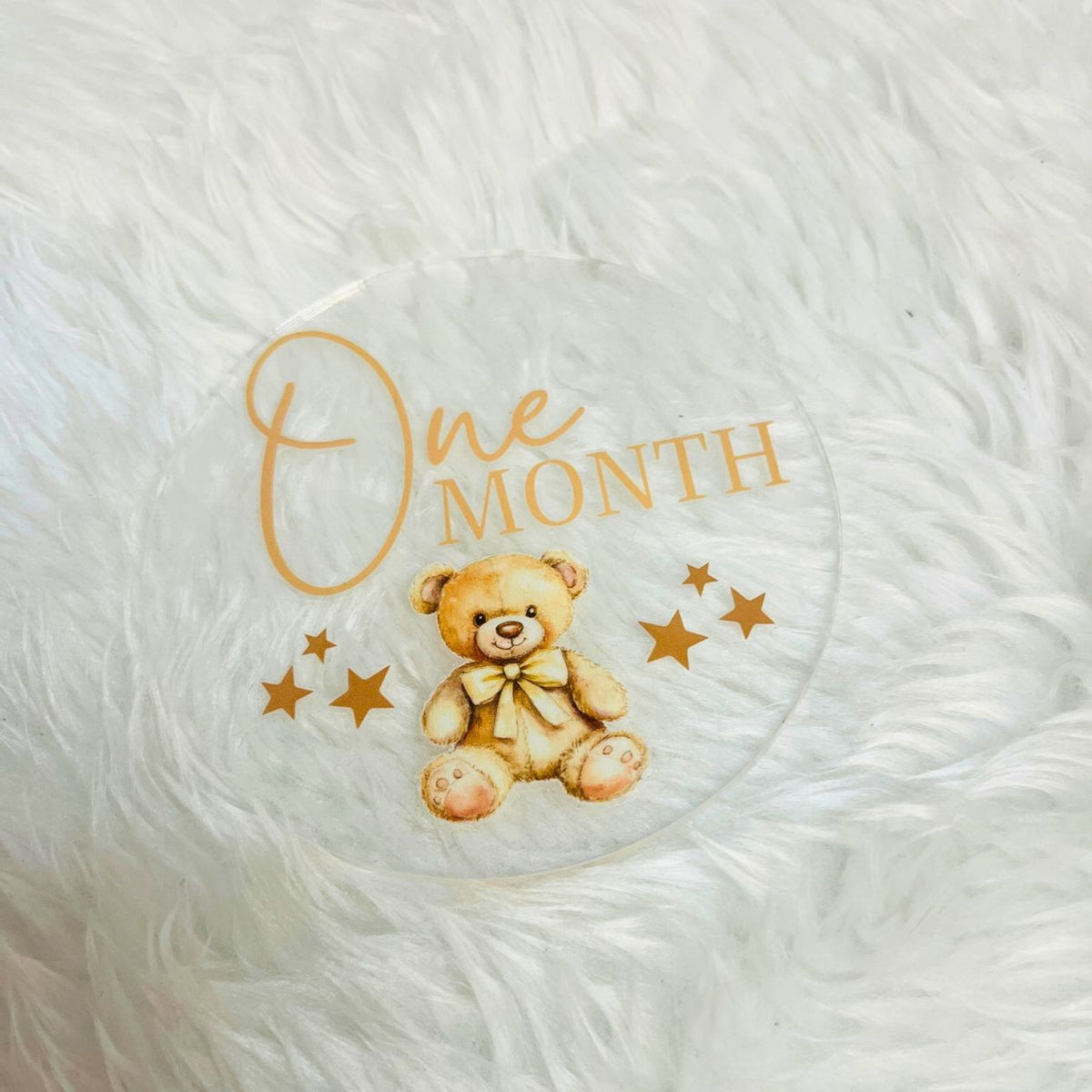 A round clear acrylic baby keepsake plaque featuring a watercolor teddy bear wearing a bow and the text 'One MONTH' in gold, displayed on a white faux fur background.