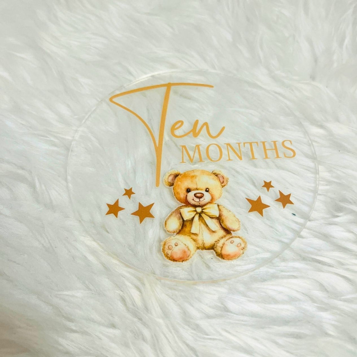 A round clear plastic or glass keepsake dish or coaster featuring a printed design of a teddy bear, gold stars, and the text 'Ten Months' in gold, displayed on a white faux fur background.