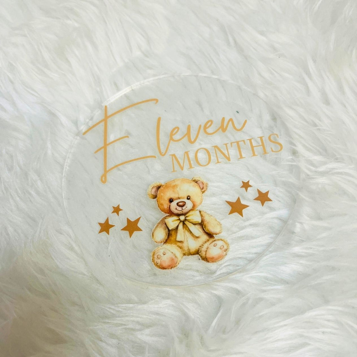 A round clear acrylic plaque or coaster featuring a watercolor teddy bear wearing a bow, surrounded by gold stars and the text 'Eleven Months' in gold script and block lettering, placed on a white faux fur surface.