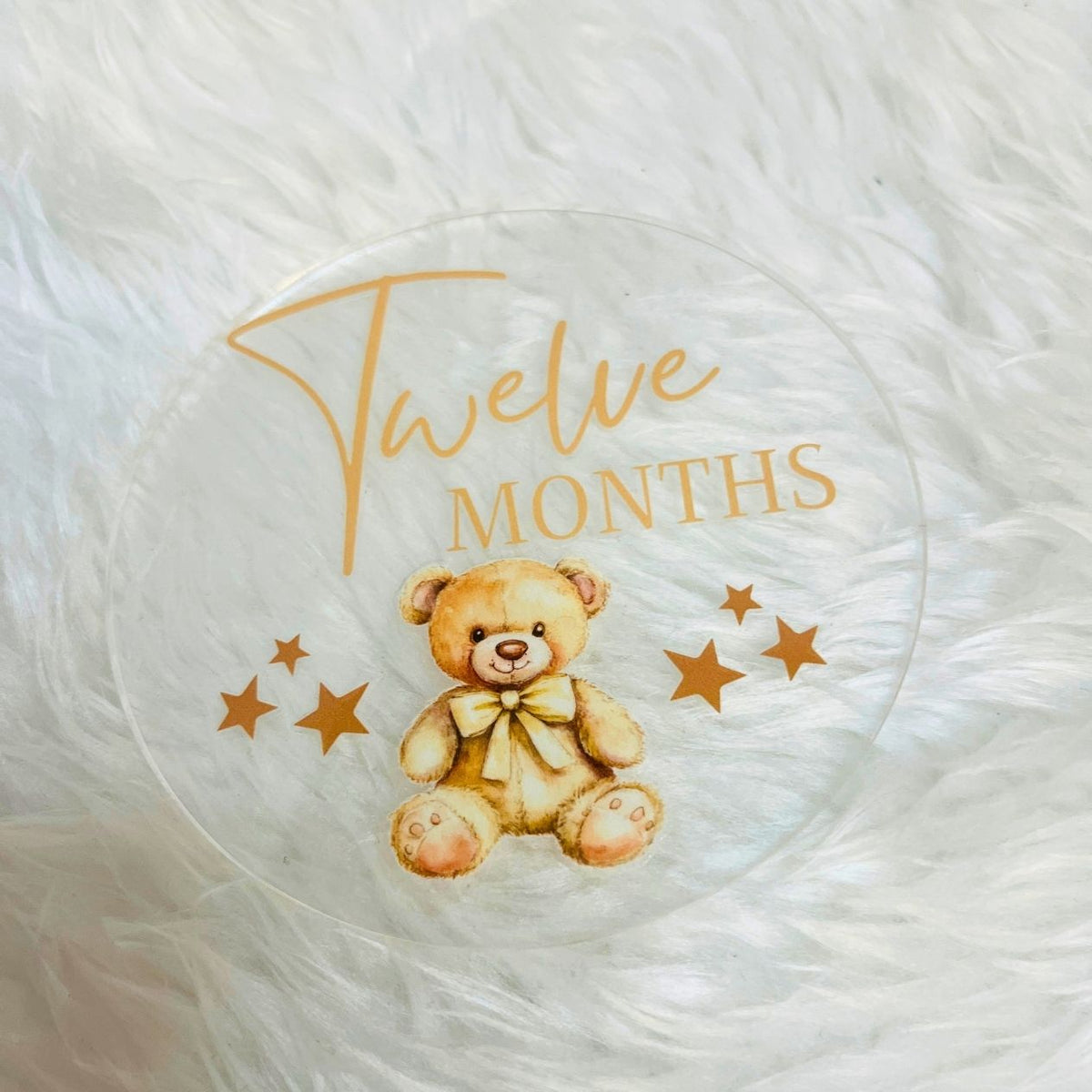 A clear round baby milestone plaque featuring a teddy bear illustration and the text 'Twelve Months' in gold, displayed on a white faux fur background.
