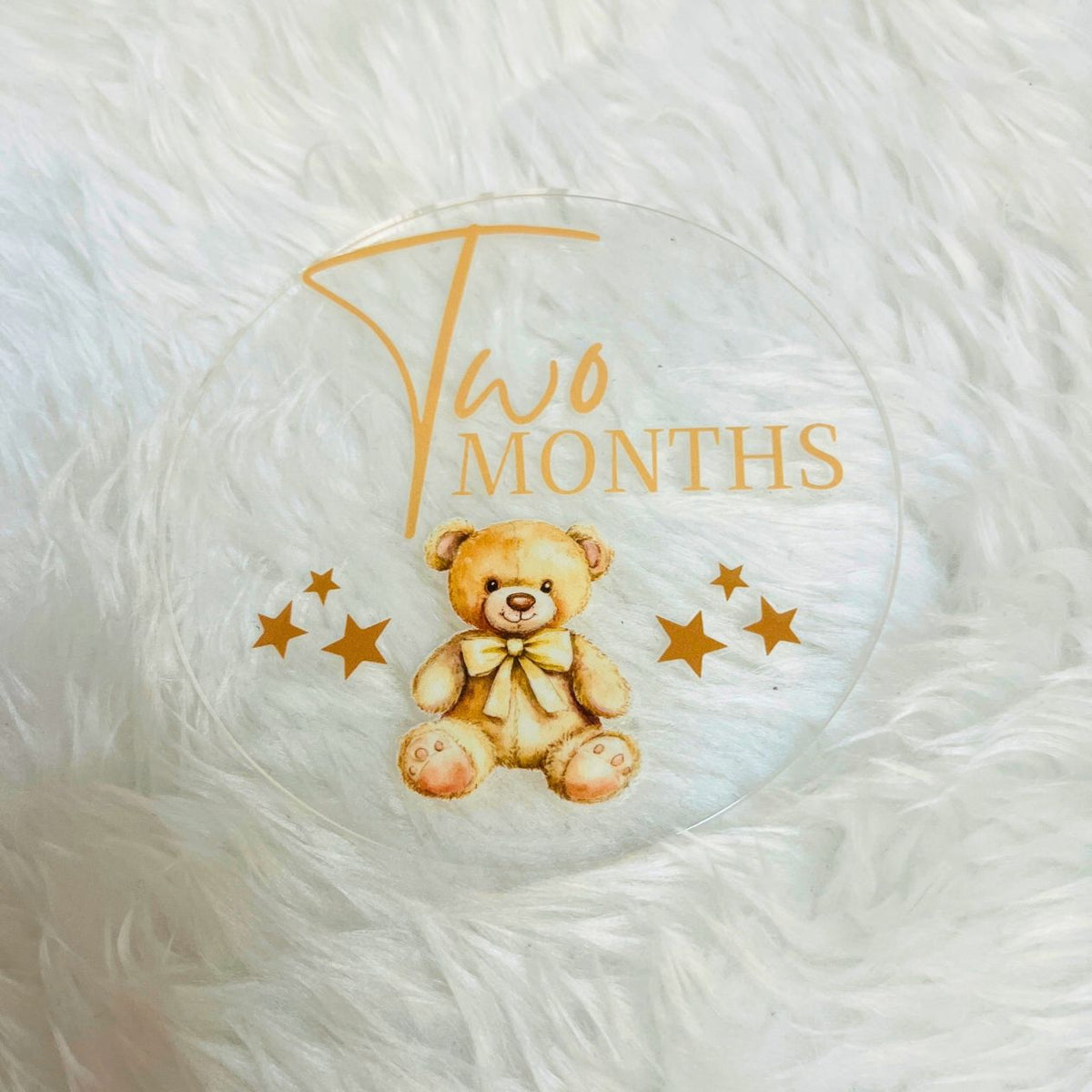 A clear round keepsake photo frame or plaque featuring a printed illustration of a teddy bear with a bow, gold stars, and the text 'Two Months' in gold, displayed on a white faux fur surface.