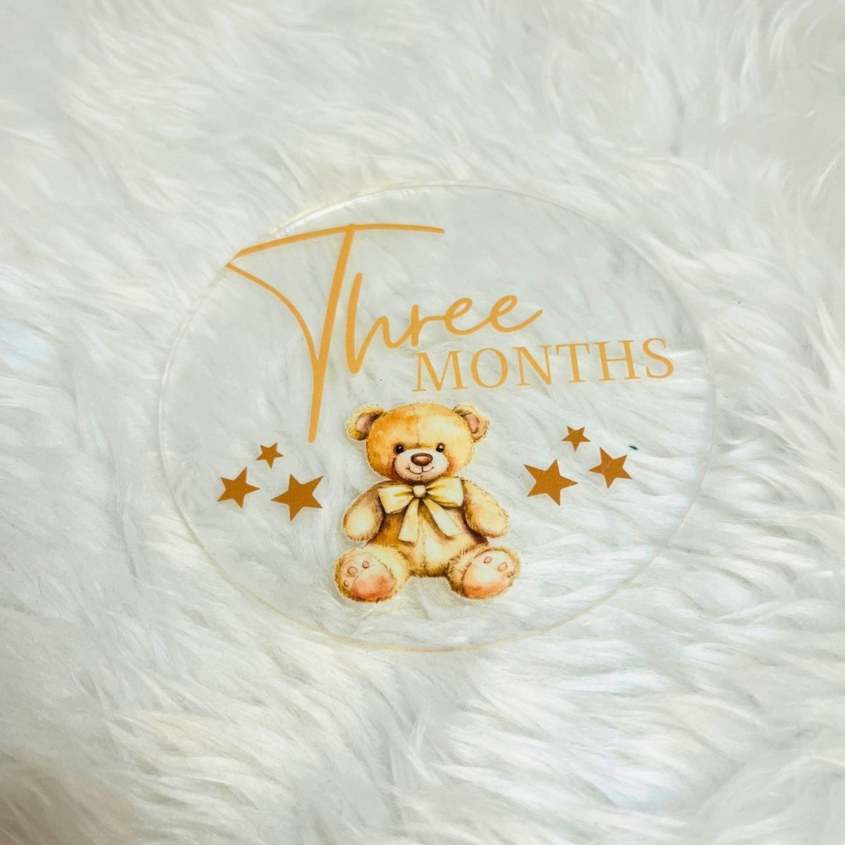 A round clear acrylic baby keepsake plaque featuring a printed teddy bear and the text 'Three Months' in gold, displayed on a white faux fur background.