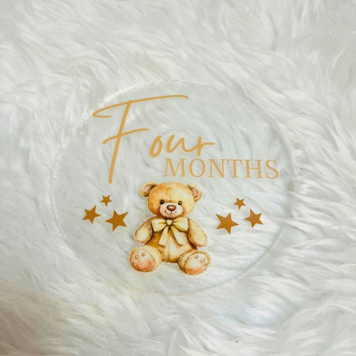 A round clear glass coaster featuring a printed illustration of a four-month-old baby teddy bear with gold text reading 'Four MONTHS' and gold stars, placed on a white faux fur surface.