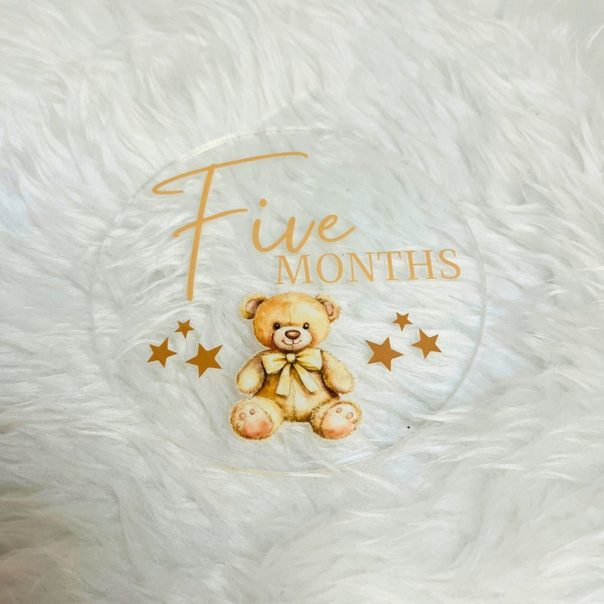 A round clear plastic baby milestone plaque celebrating 'Five Months' with a cute teddy bear illustration and gold stars, displayed on a white faux fur background.
