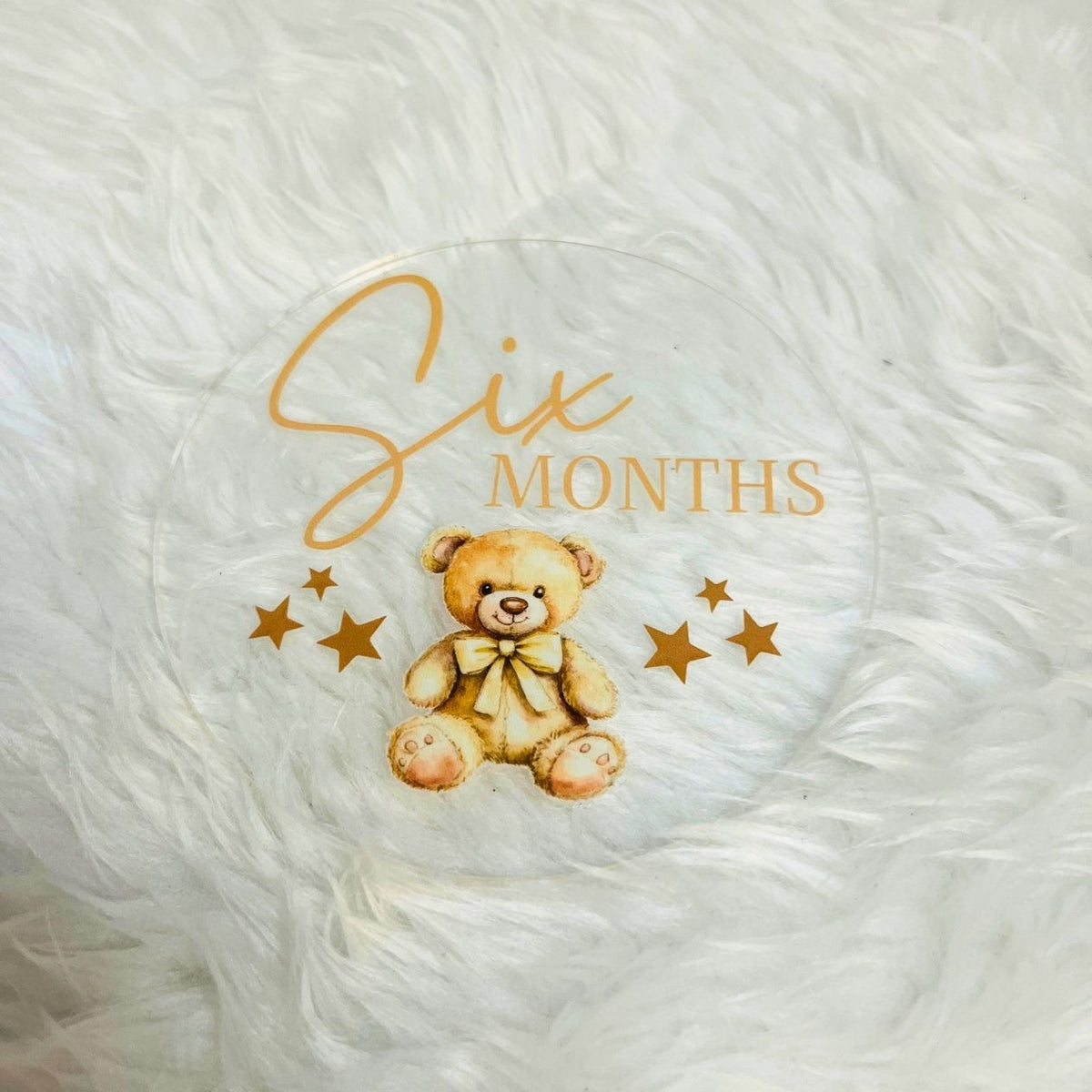 A round clear acrylic baby milestone plaque featuring a teddy bear illustration and the text 'Six Months' in gold, displayed on a white faux fur surface.
