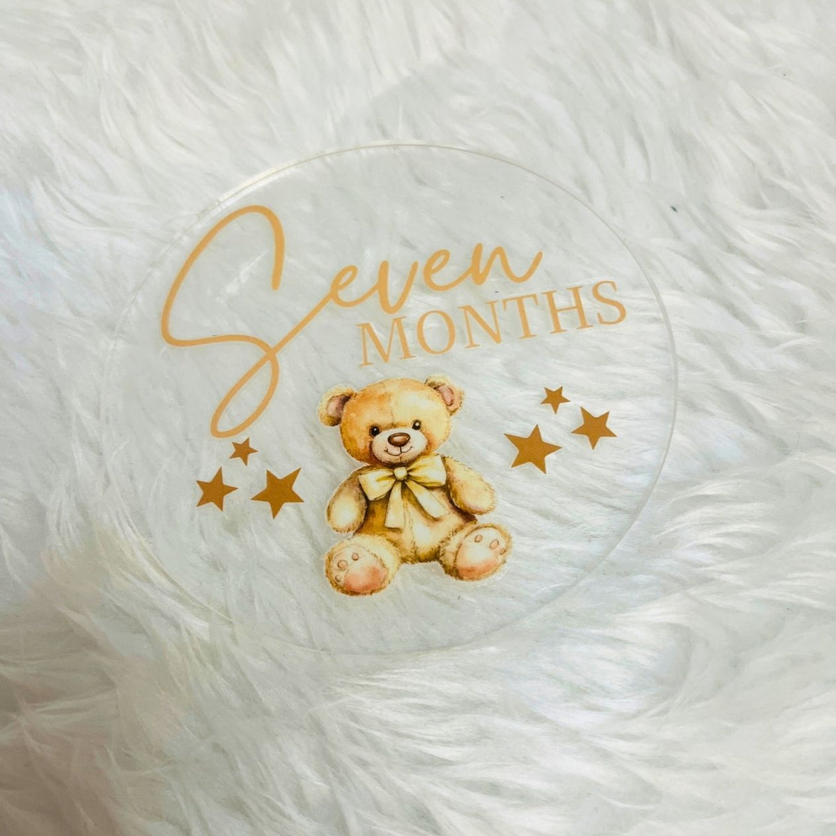 A clear round acrylic keepsake plaque featuring a watercolor teddy bear with a bow tie, gold script reading 'Seven', and gold block text reading 'MONTHS', surrounded by small gold stars. The plaque is photographed on a white faux fur surface.