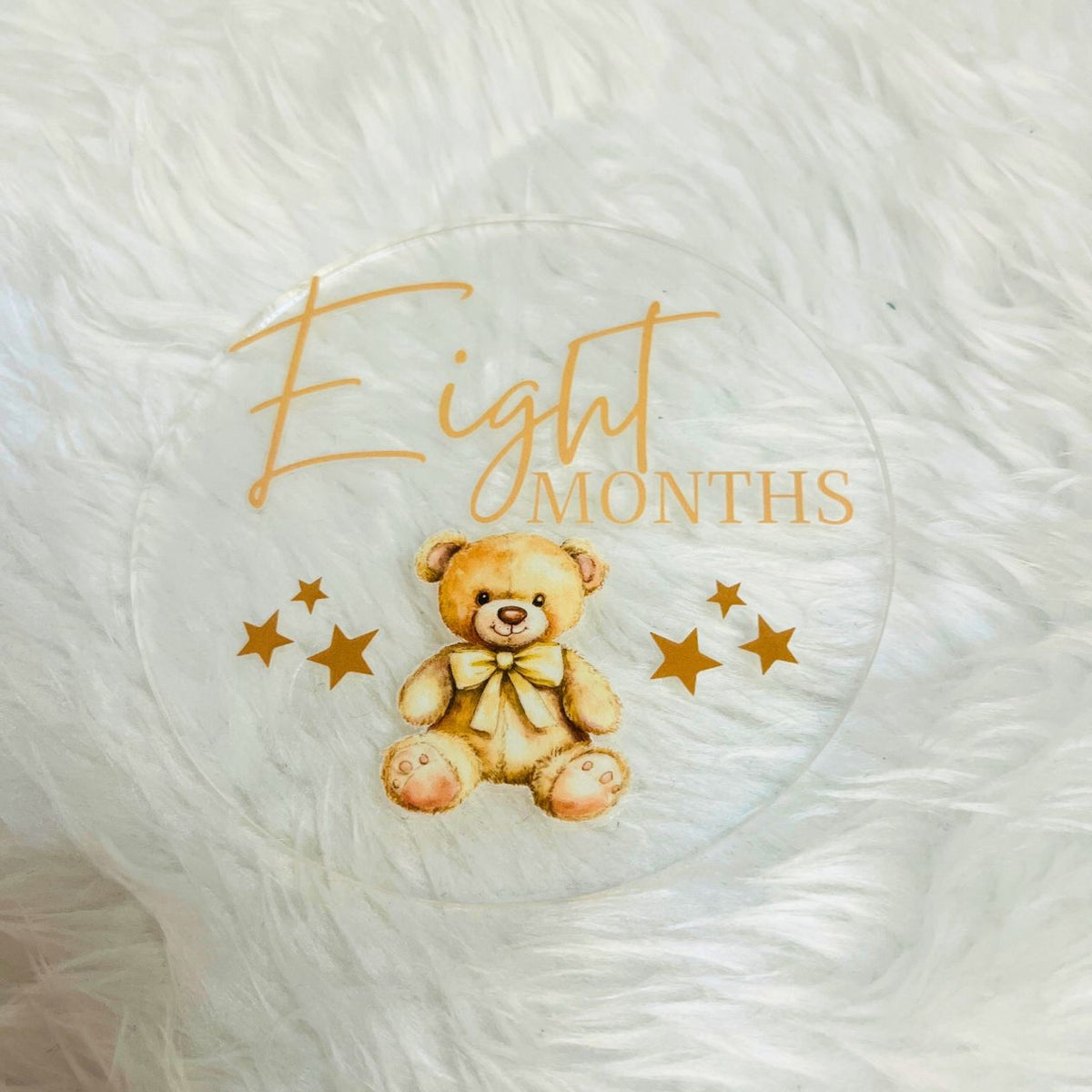 A clear round keepsake plaque featuring a watercolor teddy bear with a yellow bow, surrounded by gold stars and the text 'Eight Months' in gold, displayed on a white faux fur background.