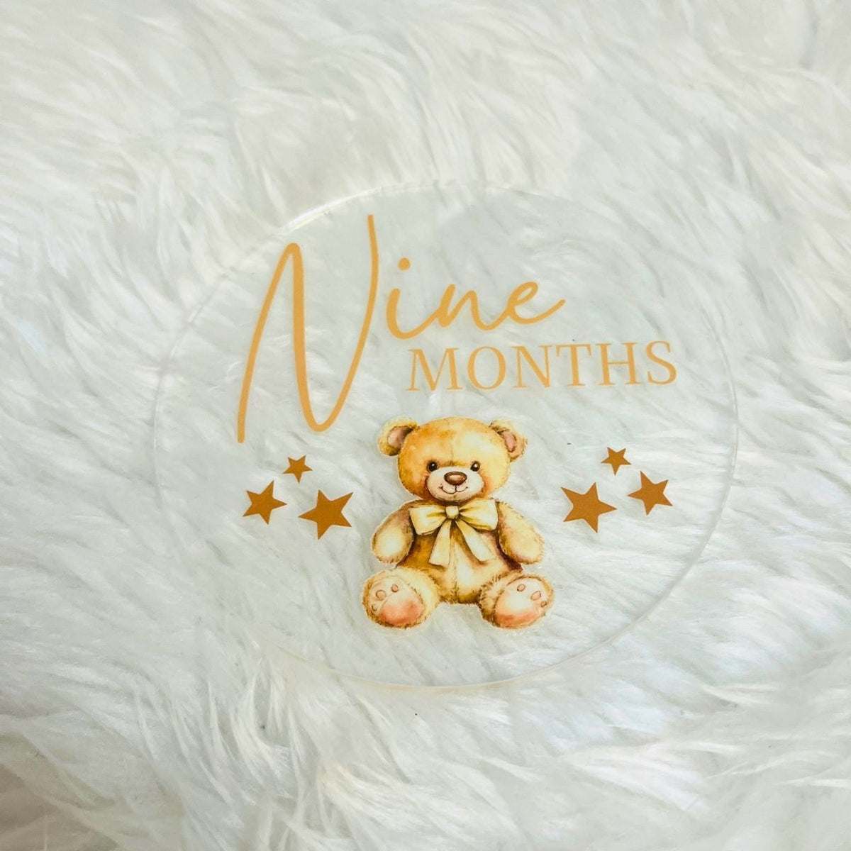 A round clear acrylic coaster featuring a printed design of a teddy bear, gold stars, and the text 'Nine Months' in gold, displayed on a white faux fur surface.