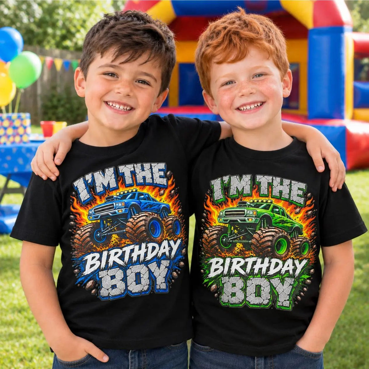 Two smiling boys standing outdoors in front of a bouncy castle, wearing matching black graphic T-shirts that read 'I'M THE BIRTHDAY BOY' with monster truck illustrations in blue and green.