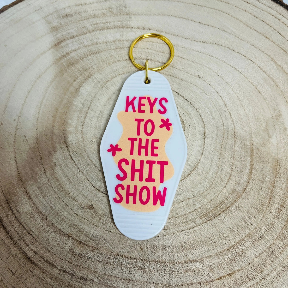 A novelty keychain featuring the text 'KEYS TO THE SHIT SHOW' in pink, layered over a light peach shape, attached to a gold split ring and photographed on a wood slice background.