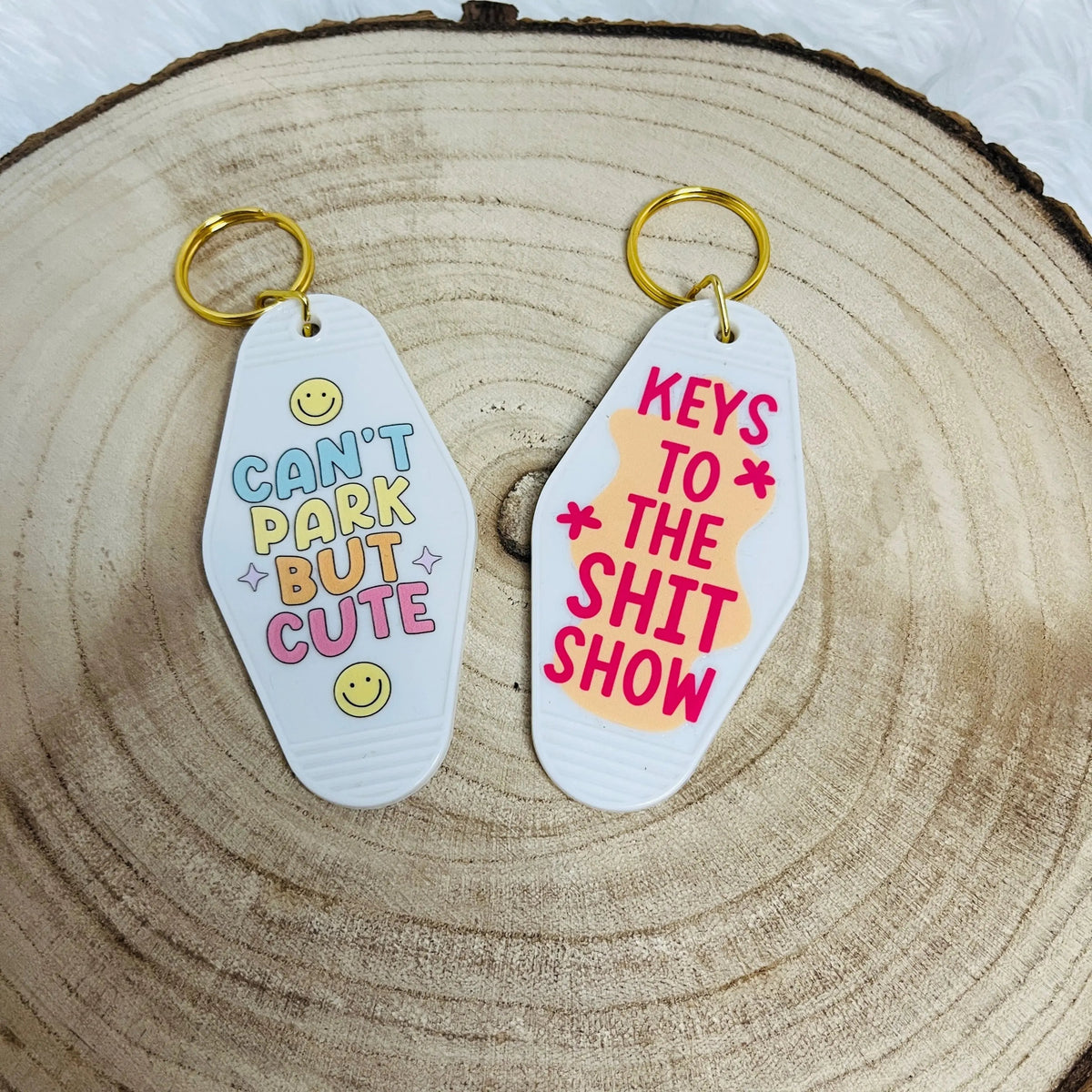 Two Motel shaped keychains displayed on a rustic wood slice background. One keychain is white with colourful text reading 'CAN'T PARK BUT CUTE', and the other is white with pink text reading 'KEYS TO THE SHIT SHOW'.