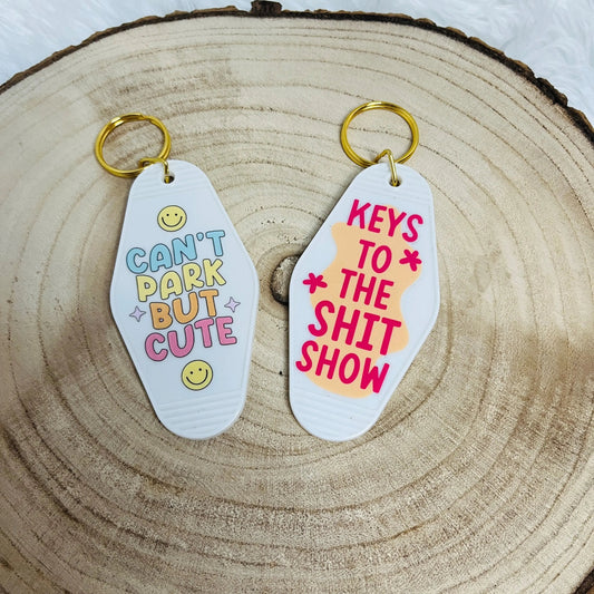 Two Motel shaped keychains displayed on a rustic wood slice background. One keychain is white with colourful text reading 'CAN'T PARK BUT CUTE', and the other is white with pink text reading 'KEYS TO THE SHIT SHOW'.