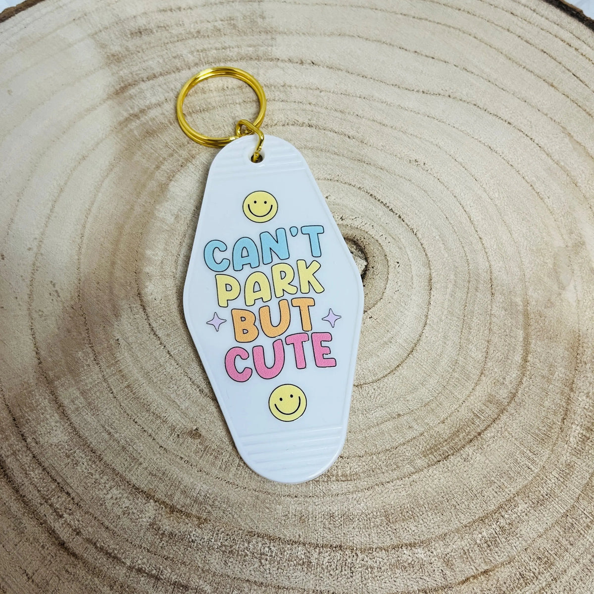 A white Motel style keychain with the text 'CAN'T PARK BUT CUTE' in pastel blue, yellow, orange, and pink lettering,  resting on a wooden slice background.