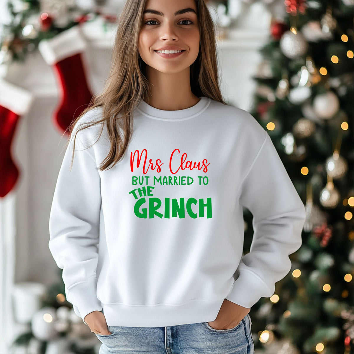 A woman wearing a white crewneck sweatshirt with a festive graphic that reads 'Mrs Claus BUT MARRIED TO THE GRINCH' in red and green lettering, posed in front of a decorated Christmas tree and stockings.