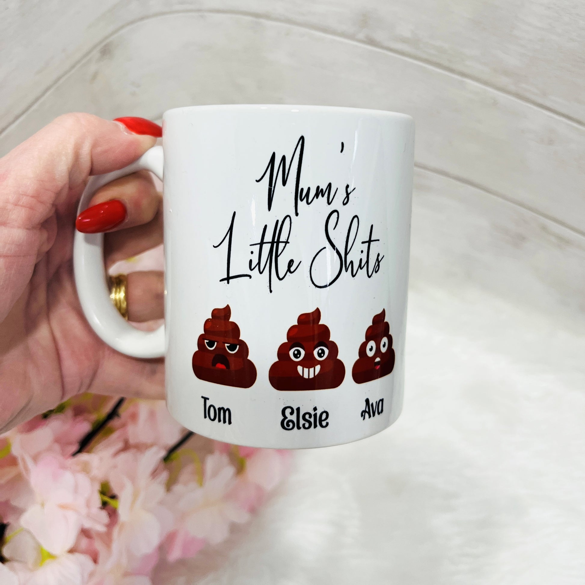 A novelty white mug with the text 'Mums little Shits' and three emojis with personalised names being held in a hand
