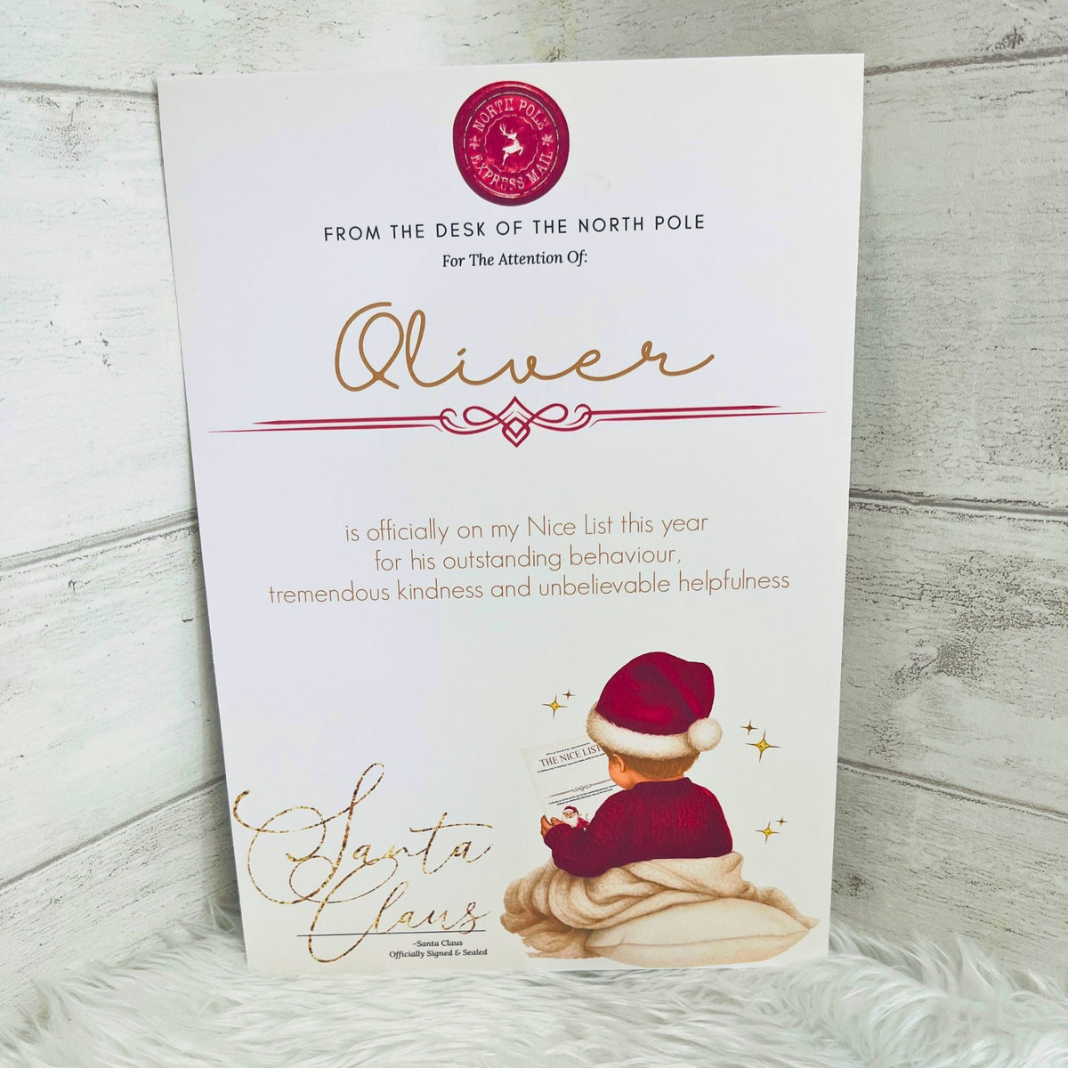 An A4 printed Nice list card addressed to 'Oliver' from the North Pole, featuring gold cursive text, a red circular logo, and an illustration of a young boy in a red jumper wearing a santa hat. The card is photographed standing upright against a light gray wood-plank background with a white faux-fur surface underneath.
