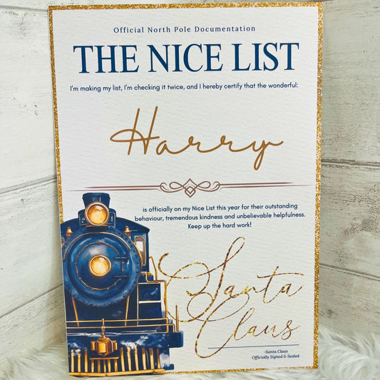 A rectangular A4 printed card with a white background and glittery gold border, featuring the text 'THE NICE LIST' and a decorative script signature reading 'Harry' above an illustration of a blue train. The sign is photographed leaning against a light wood surface with a white faux-fur surface underneath.