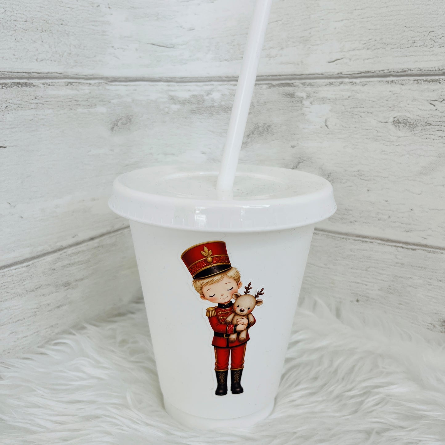 A white plastic 16oz tumbler with a matching lid and straw, featuring a cartoon nutcracker holding a reindeer printed on the side, set against a rustic white wood background on a white faux fur surface.