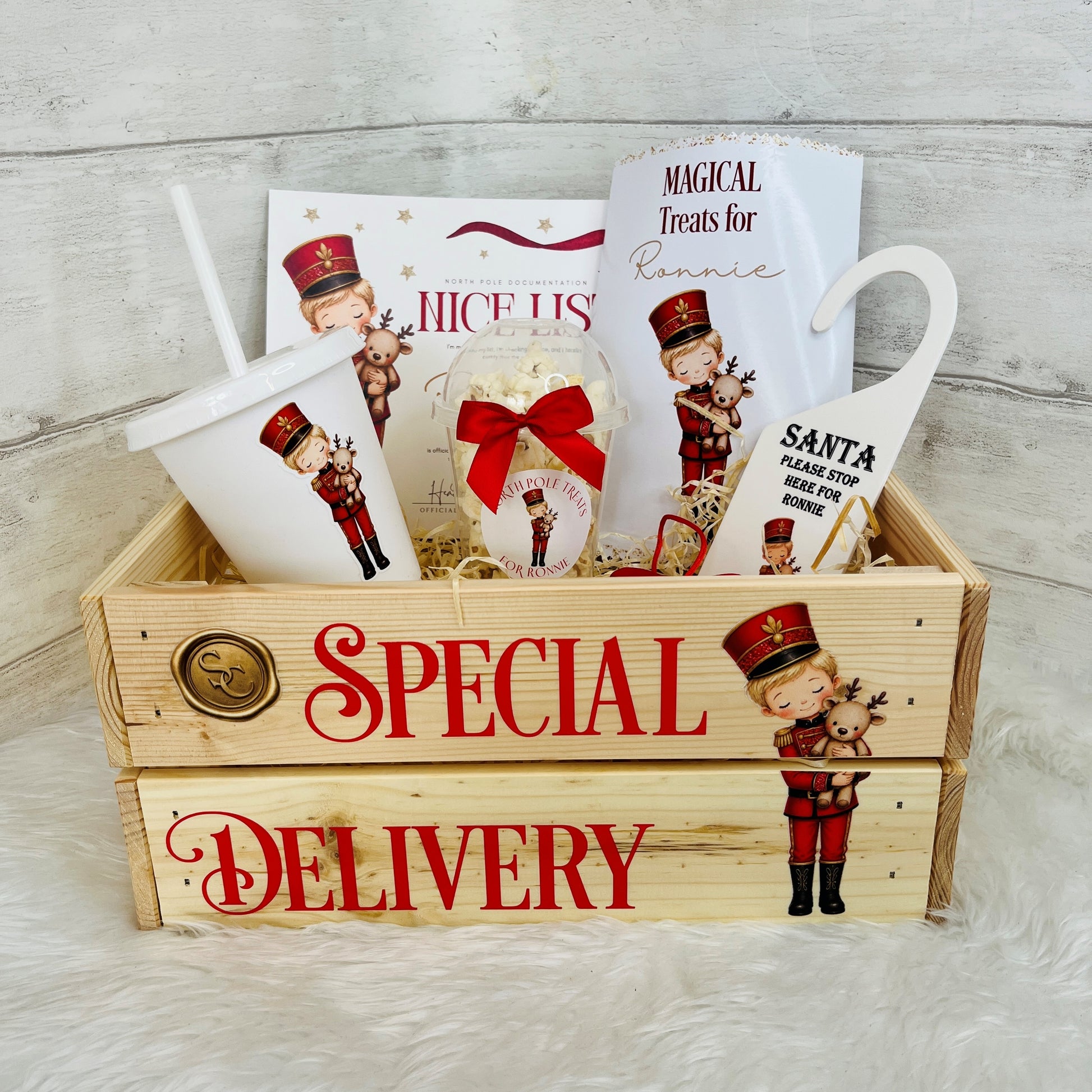 A Christmas-themed gift basket set in a wooden crate labeled 'SPECIAL DELIVERY,' filled with holiday treats including a cup, packaged snacks, a small ornament, and printed cards featuring nutcracker illustrations and the text 'MAGICAL Treats for Ronnie' and 'Santa Please Stop here for Ronnie'.