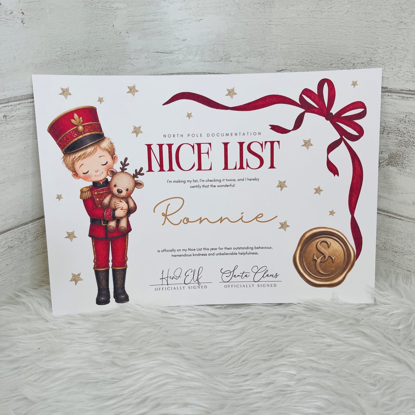 A personalised A5 Christmas 'Nice List' certificate card featuring an illustration of a nutcracker holding a teddy bear, red ribbon graphics, gold stars, and a wax seal, displayed on a white faux fur surface against a light gray wood background.