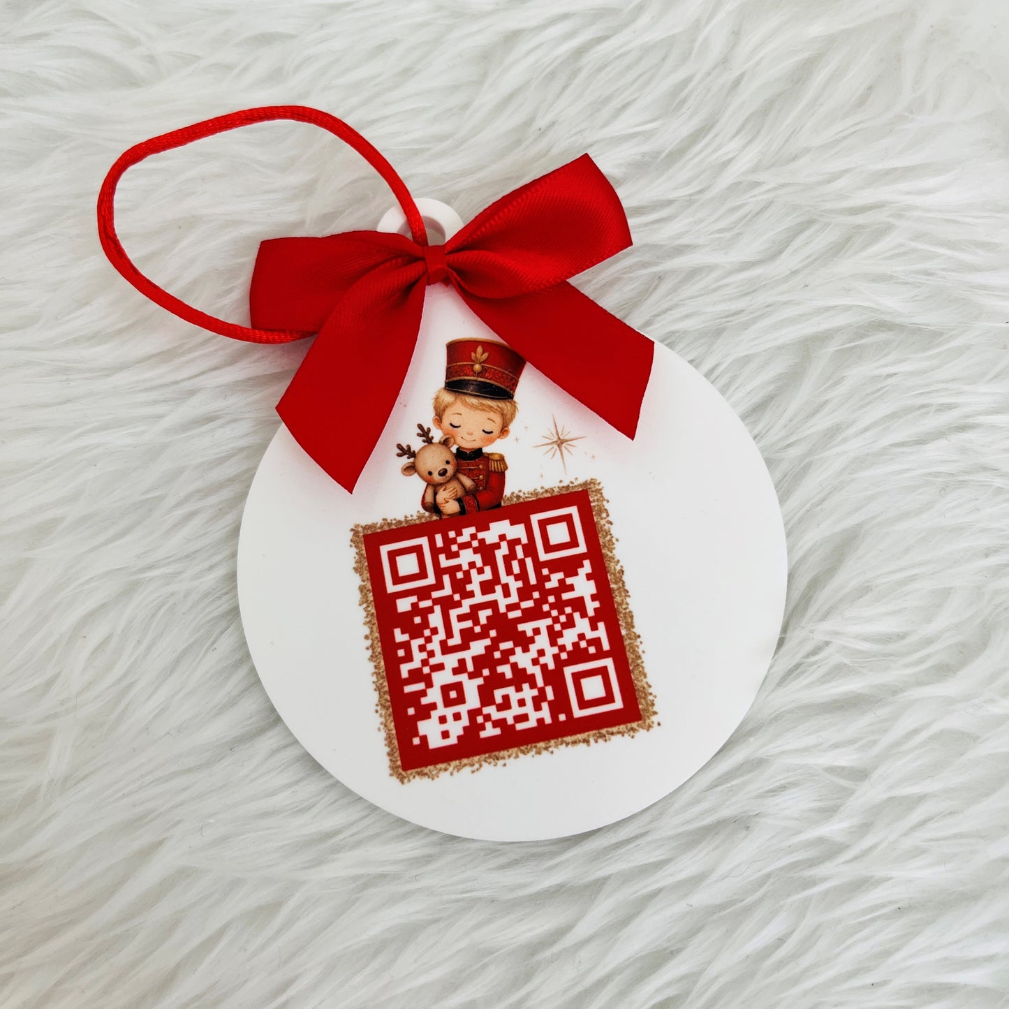 A round white Christmas ornament decorated with a red ribbon loop, a red bow, and a printed red-and-white santa tracker QR code with a small holiday character illustration, shown on a white faux-fur background.