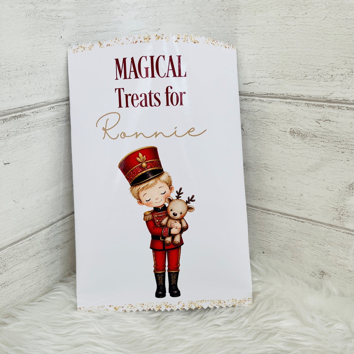 A white treat packet with scalloped gold trim, printed with the words 'MAGICAL Treats for Ronnie' and an illustration of a child in a red nutcracker uniform holding a teddy bear. The packet is photographed standing upright on a white faux-fur surface against a light wood plank background.