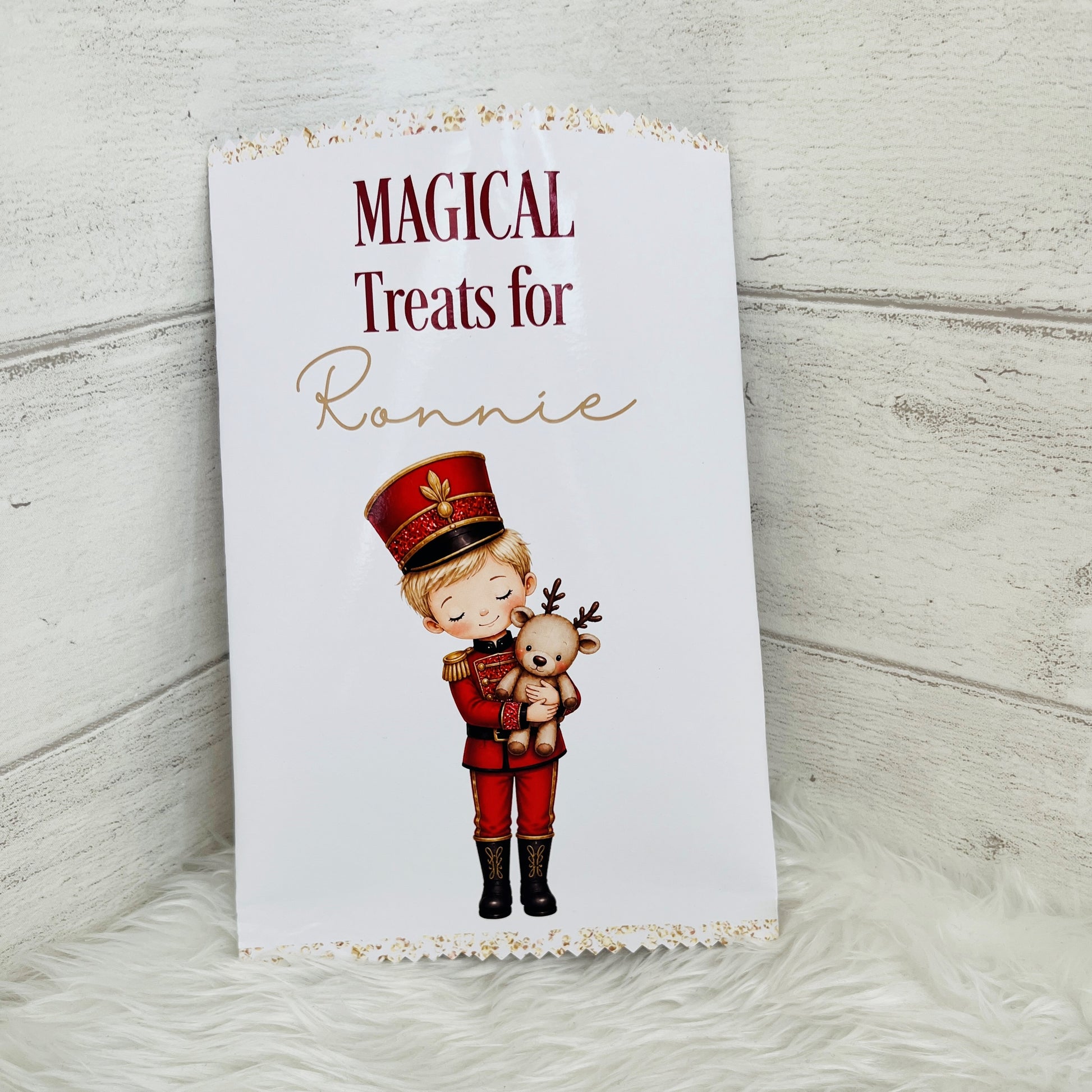 A white treat packet with scalloped gold trim, printed with the words 'MAGICAL Treats for Ronnie' and an illustration of a child in a red nutcracker uniform holding a teddy bear. The packet is photographed standing upright on a white faux-fur surface against a light wood plank background.