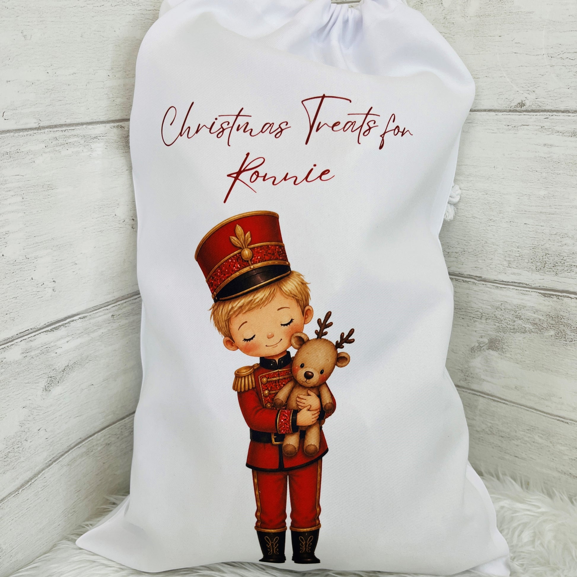 A white fabric Christmas santa sack with a drawstring top, printed with the text 'Christmas Treats for Ronnie' in dark red script and an illustration of a child soldier nutcracker holding a teddy bear. The bag is displayed on a rustic white-washed wooden surface.
