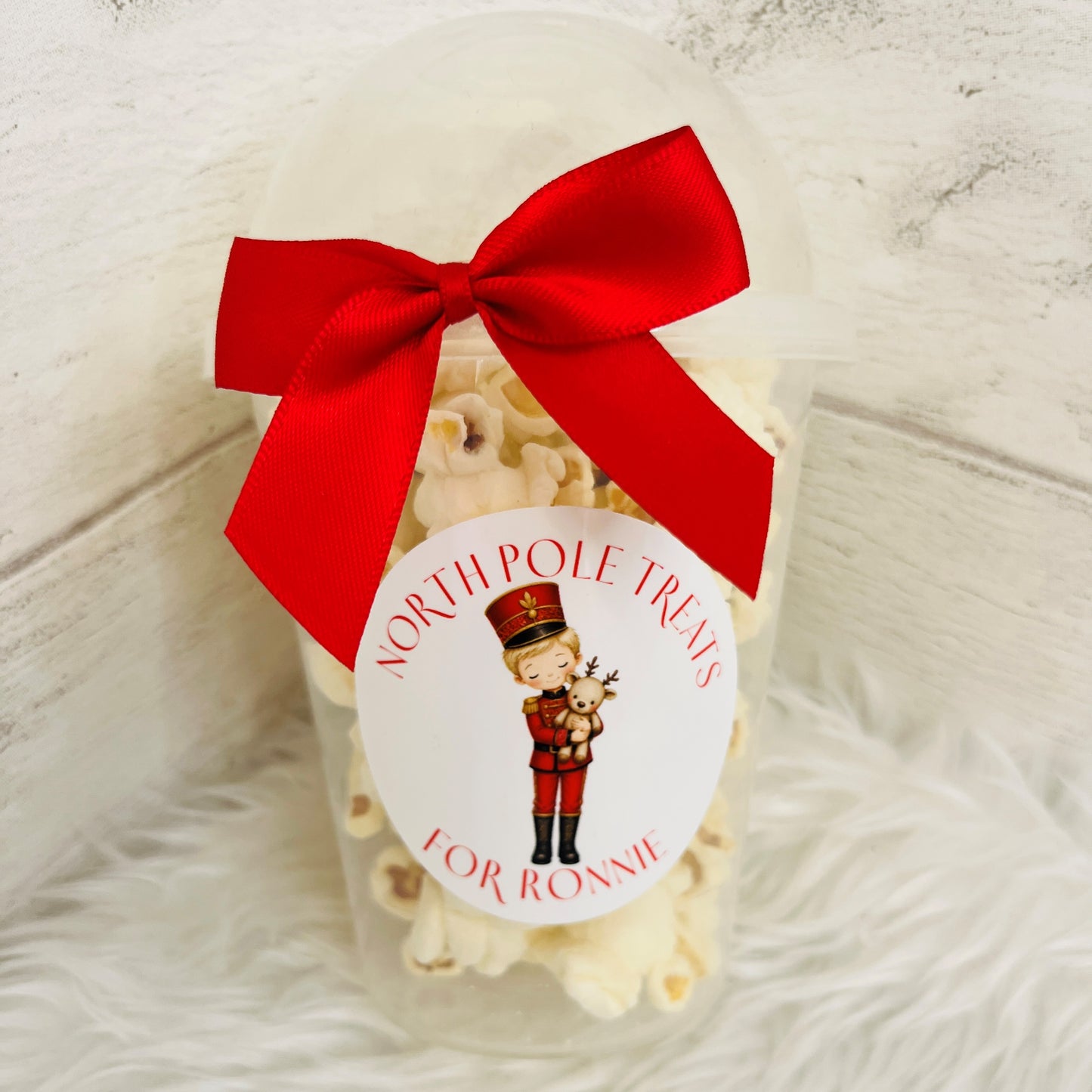 A clear plastic tub filled with popcorn, tied with a large red ribbon and a round white sticker that reads 'NORTH POLE TREATS FOR RONNIE' with a nutcracker illustration, set on a white faux-fur surface against a light wood background.