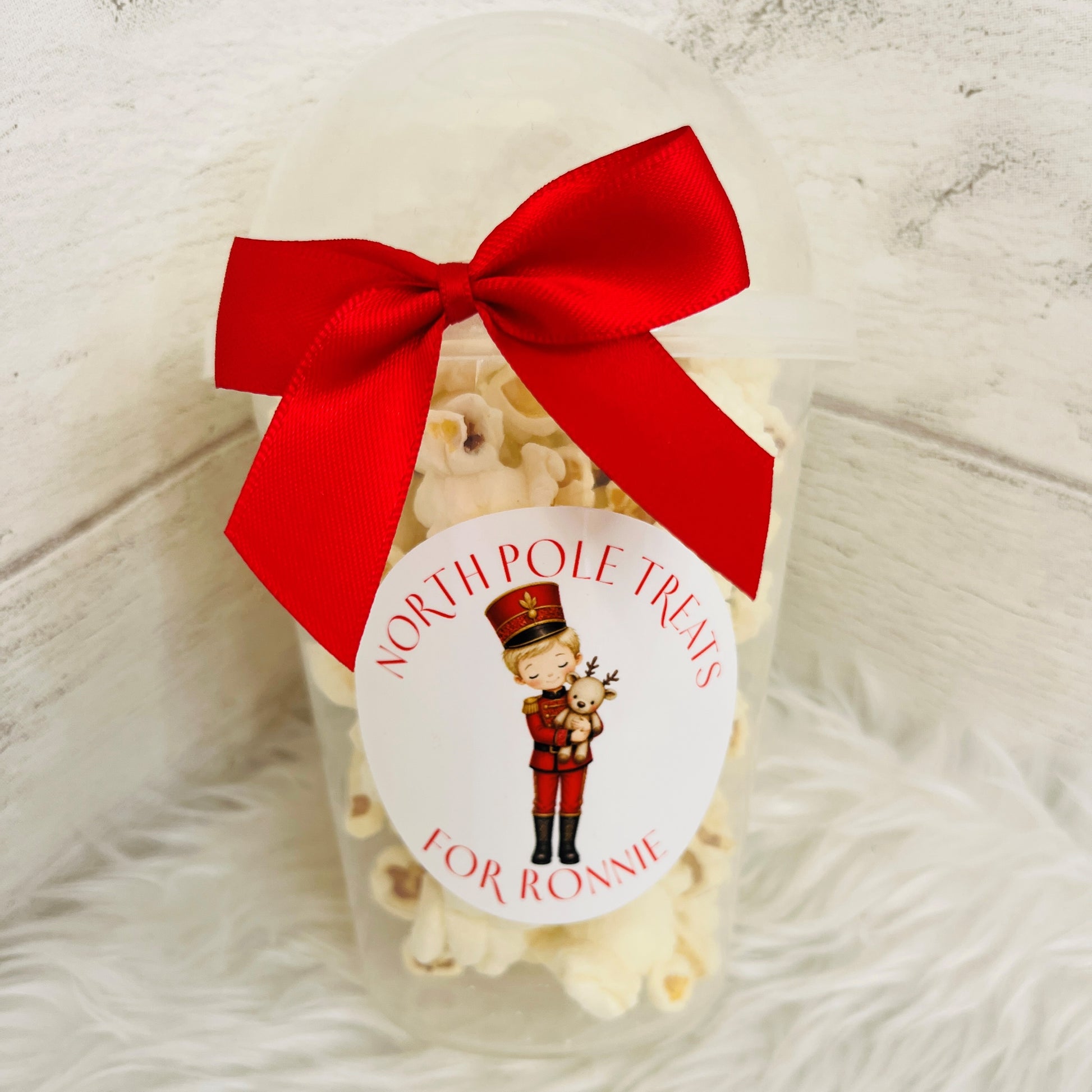 A clear plastic tub filled with popcorn, tied with a large red ribbon and a round white sticker that reads 'NORTH POLE TREATS FOR RONNIE' with a nutcracker illustration, set on a white faux-fur surface against a light wood background.