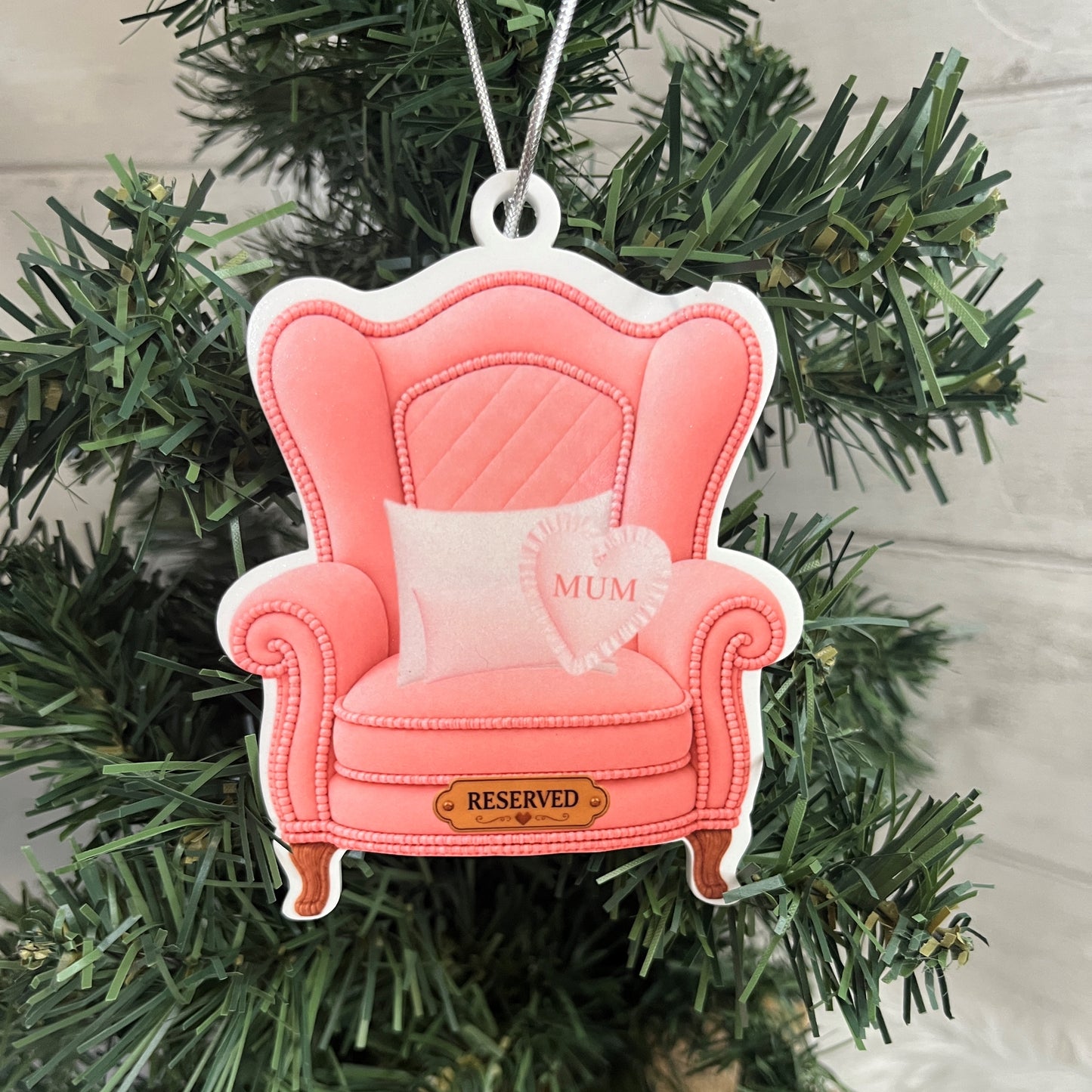 A pink armchair-shaped Christmas ornament hanging on a green Christmas tree. The ornament features a quilted backrest, rolled arms, small wooden-style legs, a white pillow with a pink heart that says 'MUM', and a small gold plaque at the front reading 'RESERVED'.