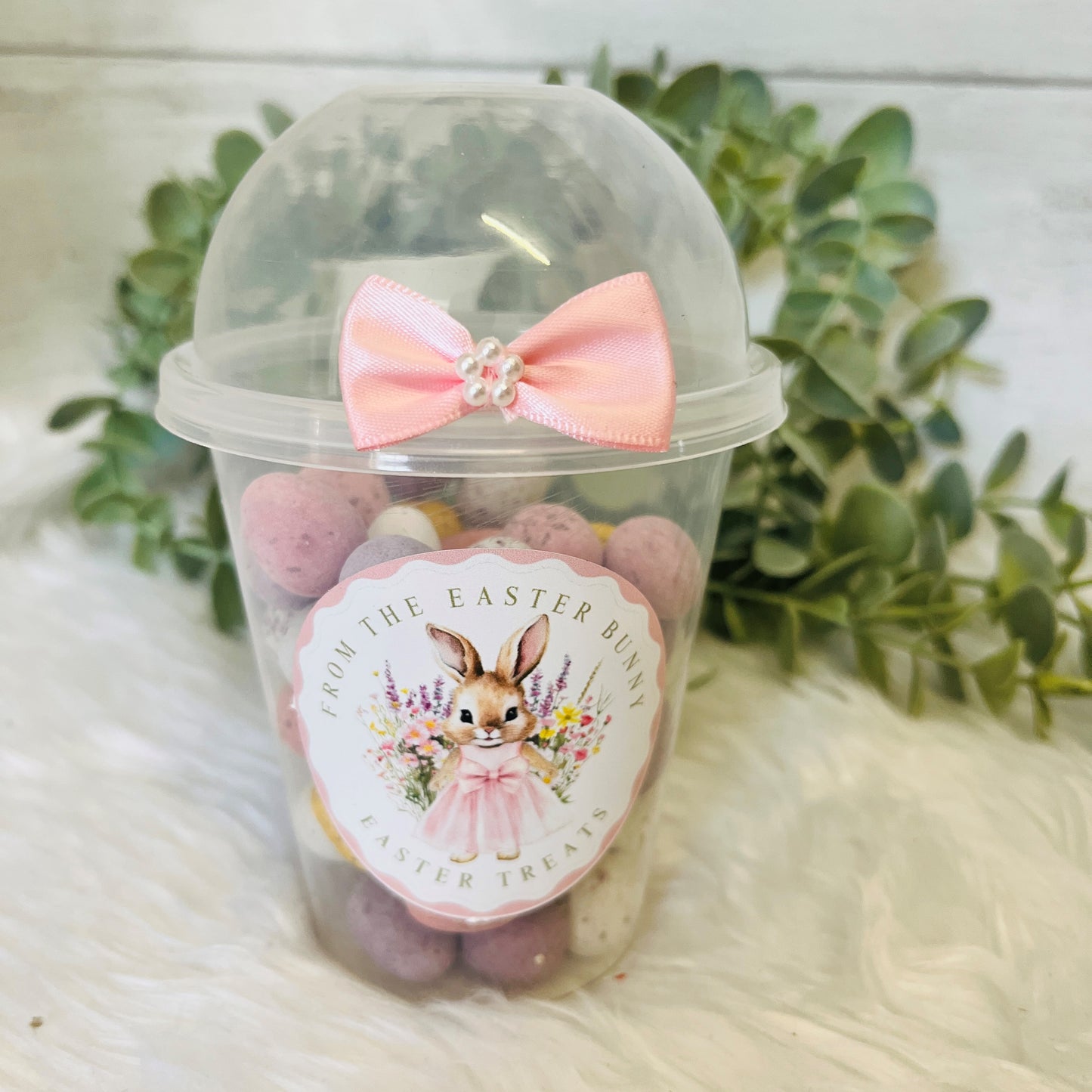 An Easter Dome Snack Pot featuring a plastic cup with a pink bunny wreath design and a pink bow on the lid, filled with various small snacks.