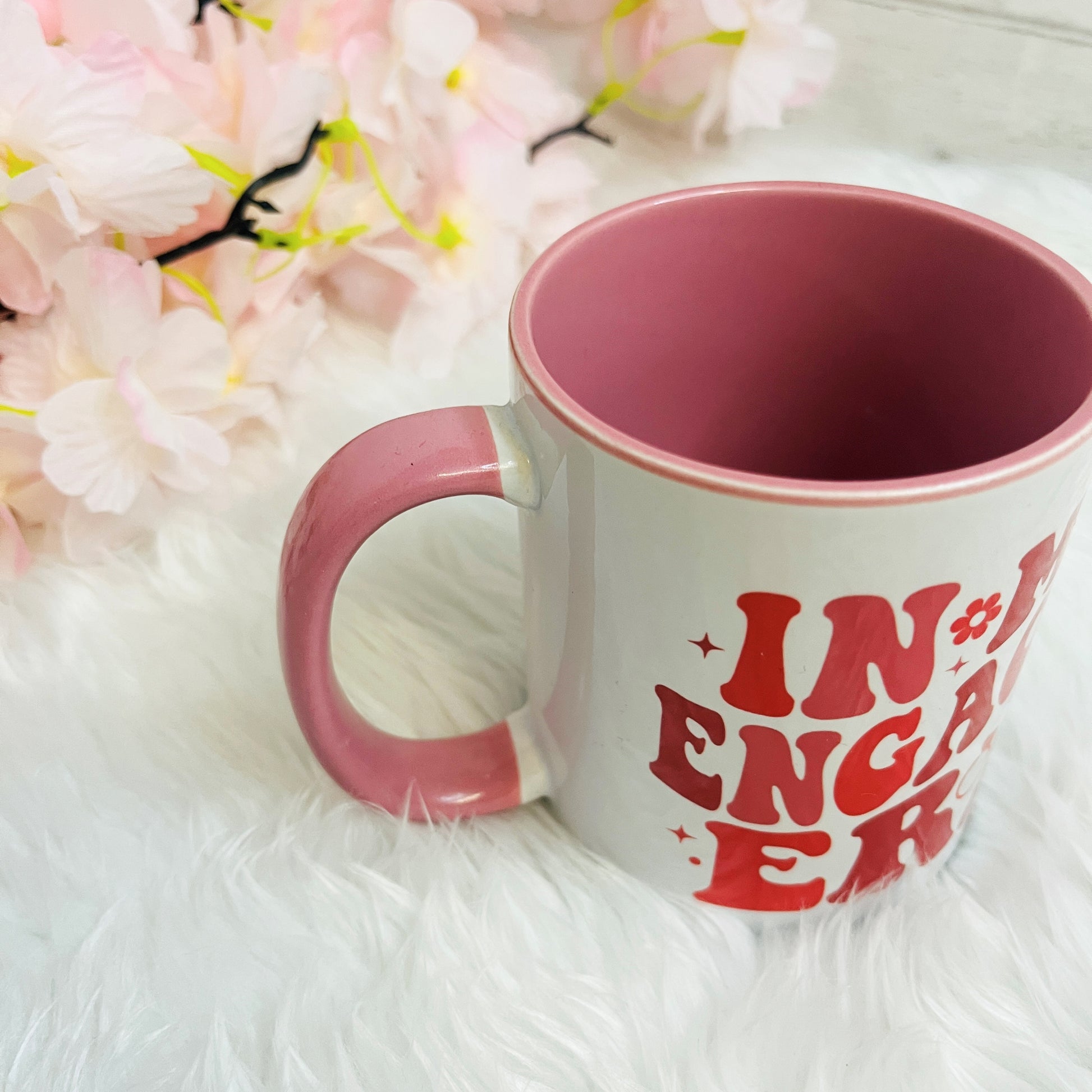 Pink mug with pink text on a white surface with flowers in the background