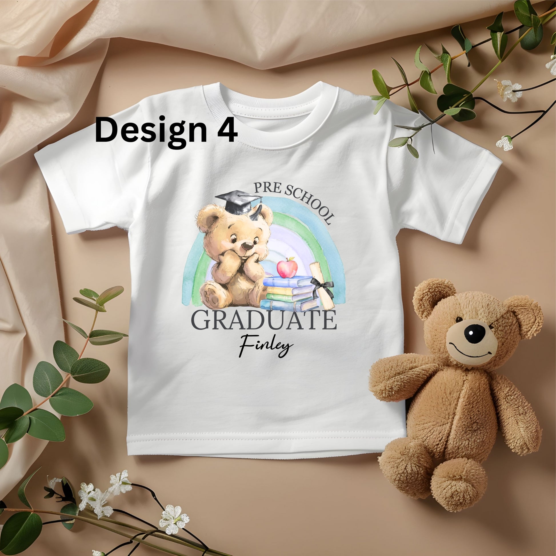 White t-shirt with teddy bear design and text, placed on a beige fabric background with a teddy bear and greenery.