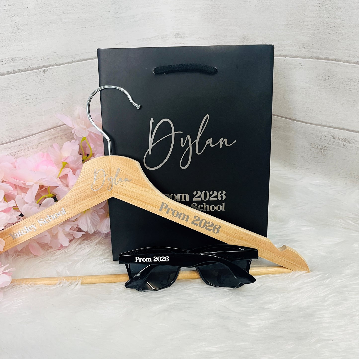 Personalized wooden hanger with 'Prom 2026' text, sunglasses, and black gift bag on a light background.