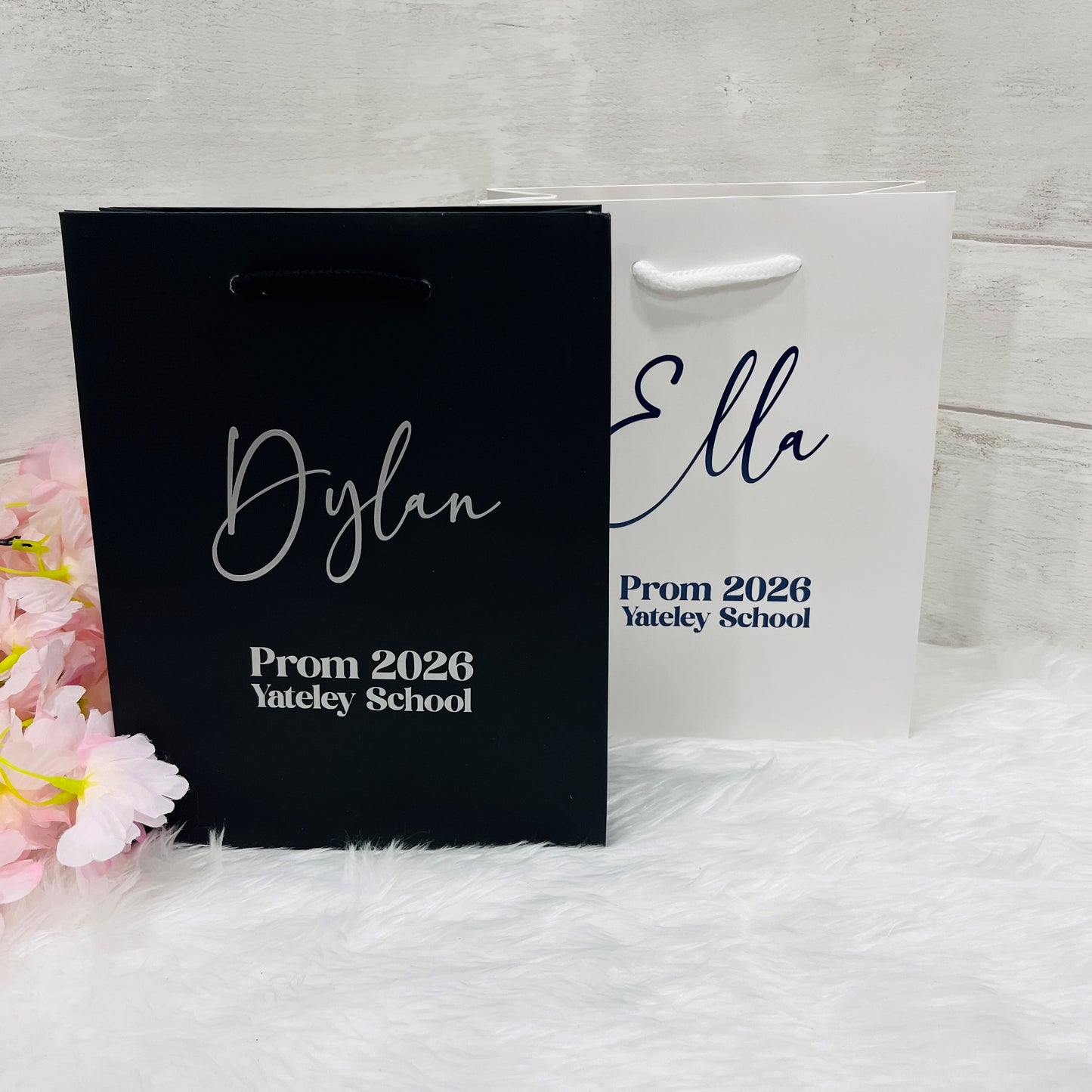 Two personalized gift bags with names and event details on a white surface with pink flowers.