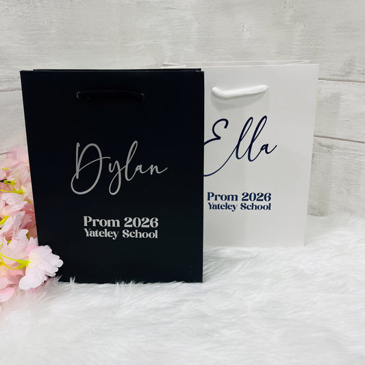 Two personalized gift bags with names and event details on a white surface with pink flowers.