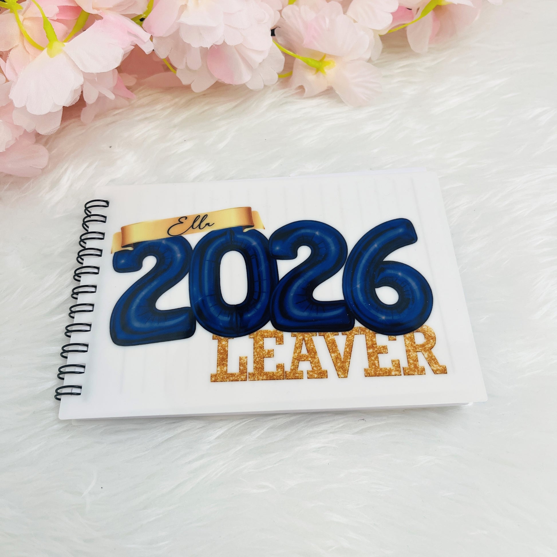 Notebook with '2026 Leaver' text in a navy design on a white surface with pink flowers in the background