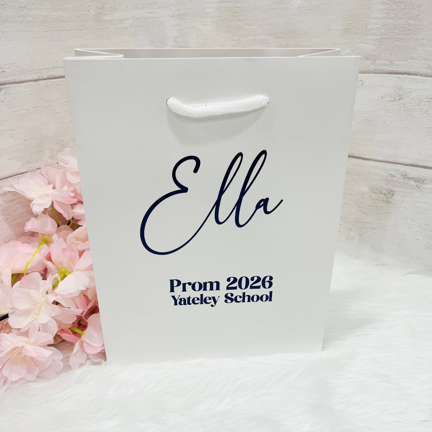 White gift bag with 'Ella Prom 2026 Yateley School' text on a white surface with pink flowers.
