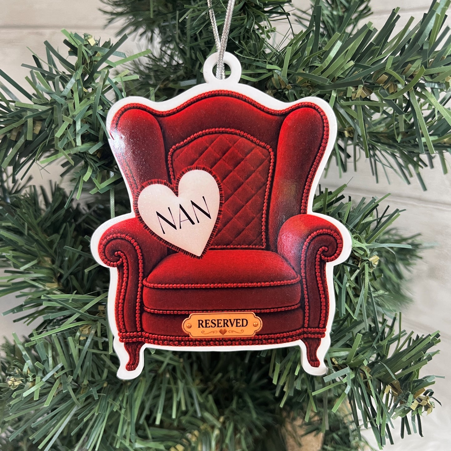 A red wingback armchair Christmas ornament hanging on a green Christmas tree. The ornament features a quilted red armchair design with a white heart-shaped cushion printed with the word 'NAN' and a small gold-colored plaque at the bottom that reads 'RESERVED'.