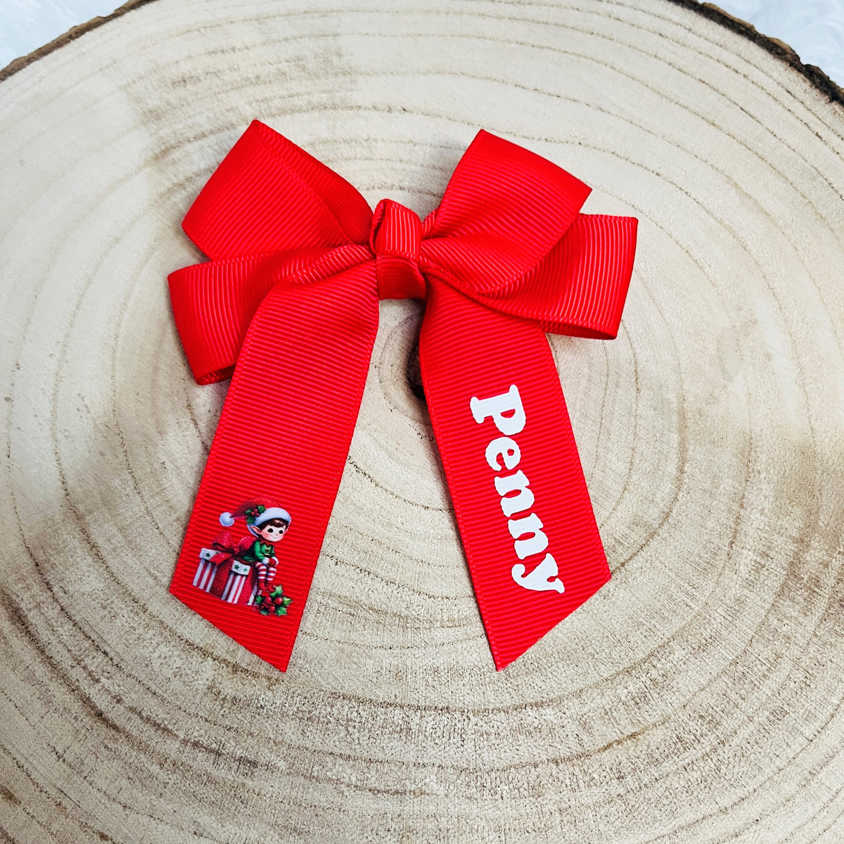 A bright red grosgrain ribbon bow with two tails, one printed with the name 'Penny' in white and the other featuring a small christmas elf graphic, displayed on a circular wood slice background.