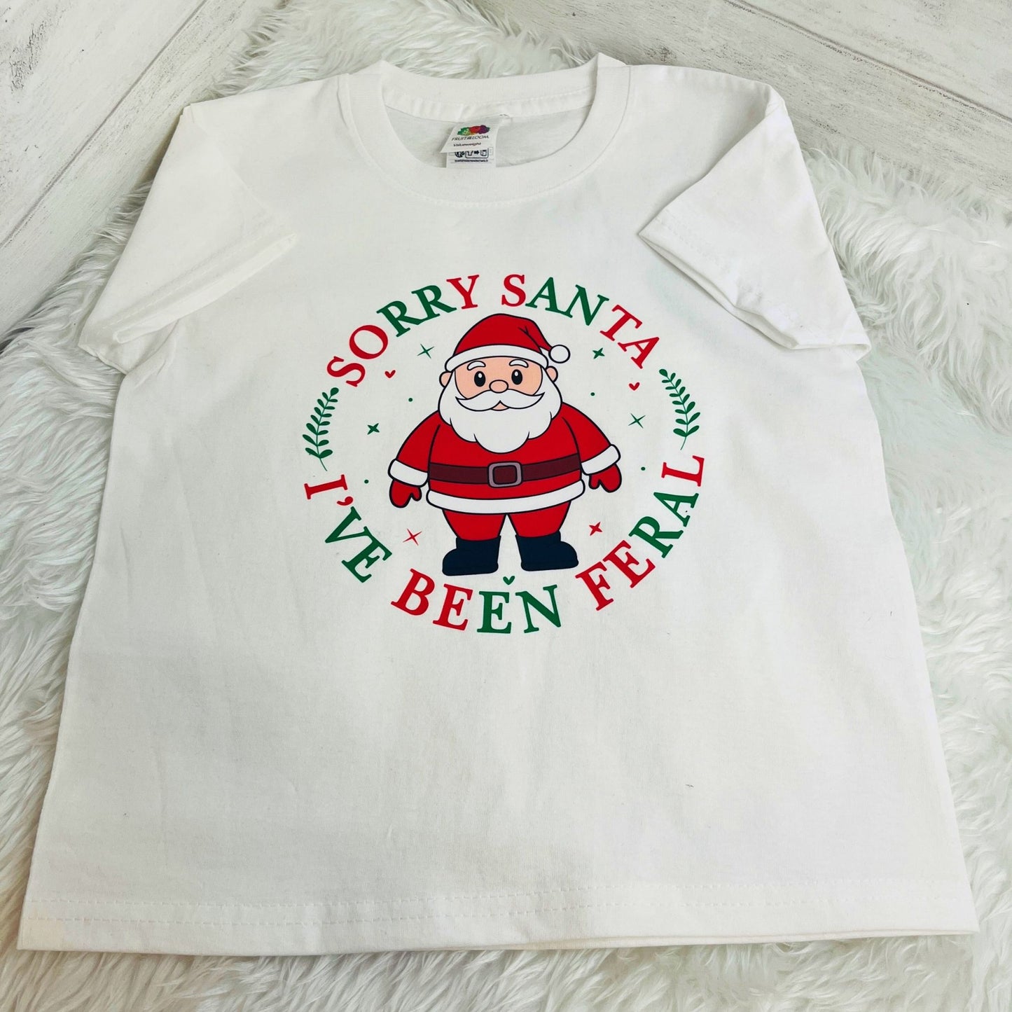 A white short-sleeve t-shirt laid flat on a white faux fur surface, featuring a circular graphic of Santa Claus with the text 'SORRY SANTA I'VE BEEN FERAL' in red and green lettering.