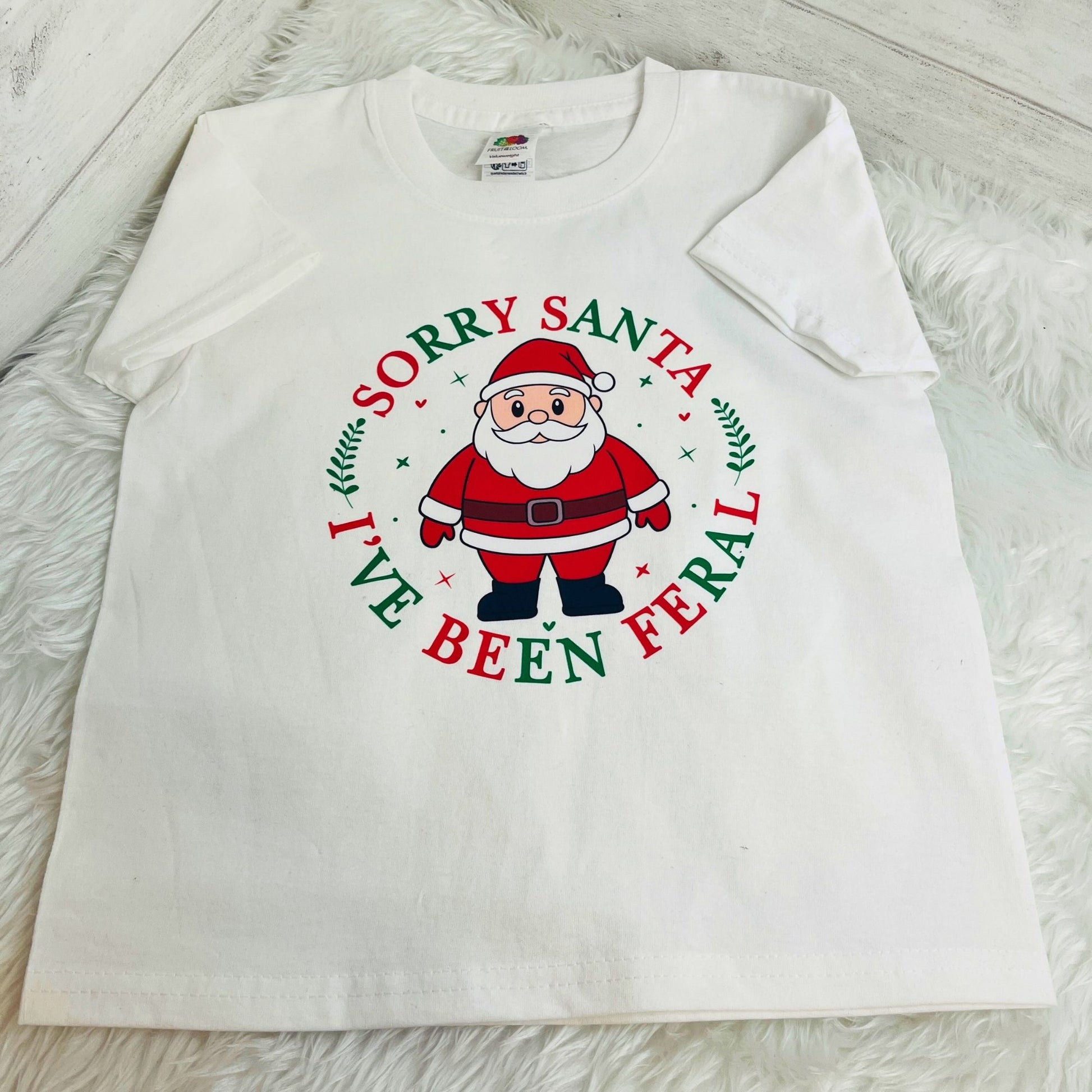 A white short-sleeve t-shirt laid flat on a white faux fur surface, featuring a circular graphic of Santa Claus with the text 'SORRY SANTA I'VE BEEN FERAL' in red and green lettering.
